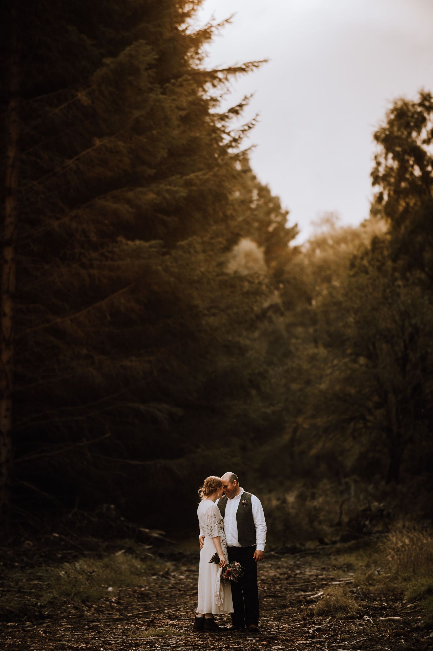 Romantic woodland wedding photo with couple silhouetted against dark forest backdrop.