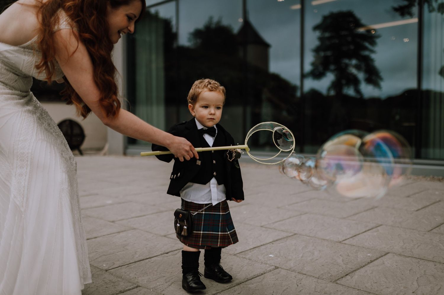Young child in kilt plays with bubbles during wedding celebration.