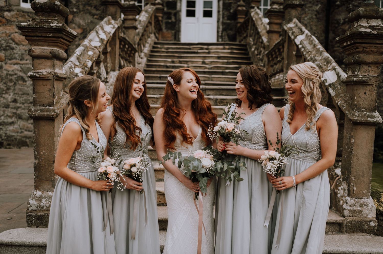Bridesmaids in pale grey dresses gather on historic stone steps during wedding ceremony.