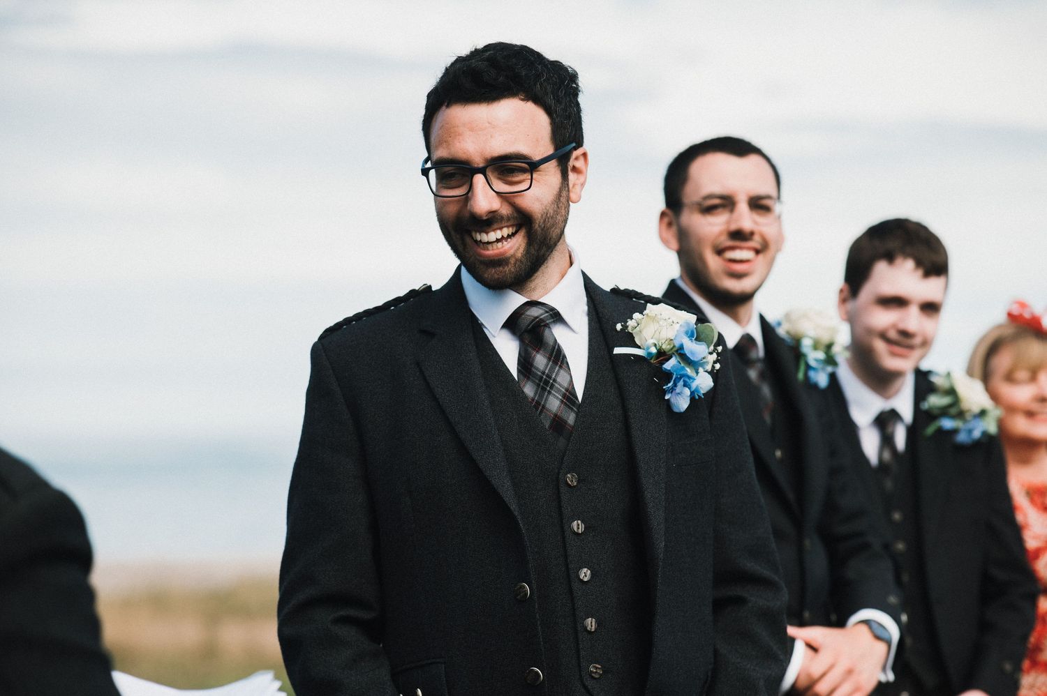 Wedding party members smile and laugh during an outdoor ceremony against a cloudy sky.