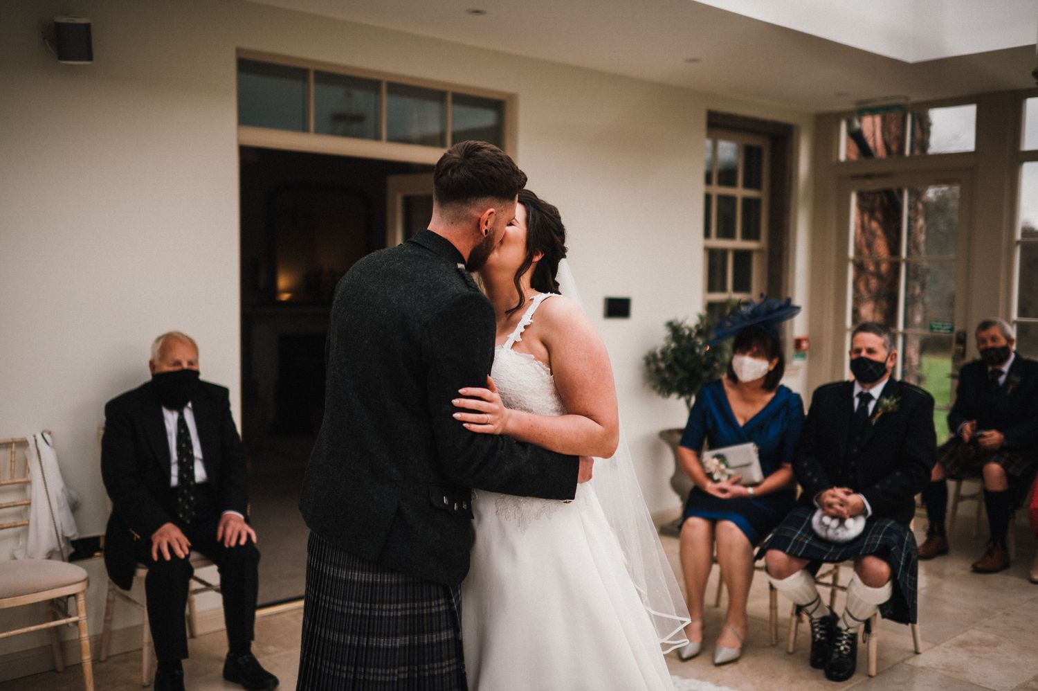 A tender moment during a Scottish wedding ceremony as guests look on in a bright indoor venue.