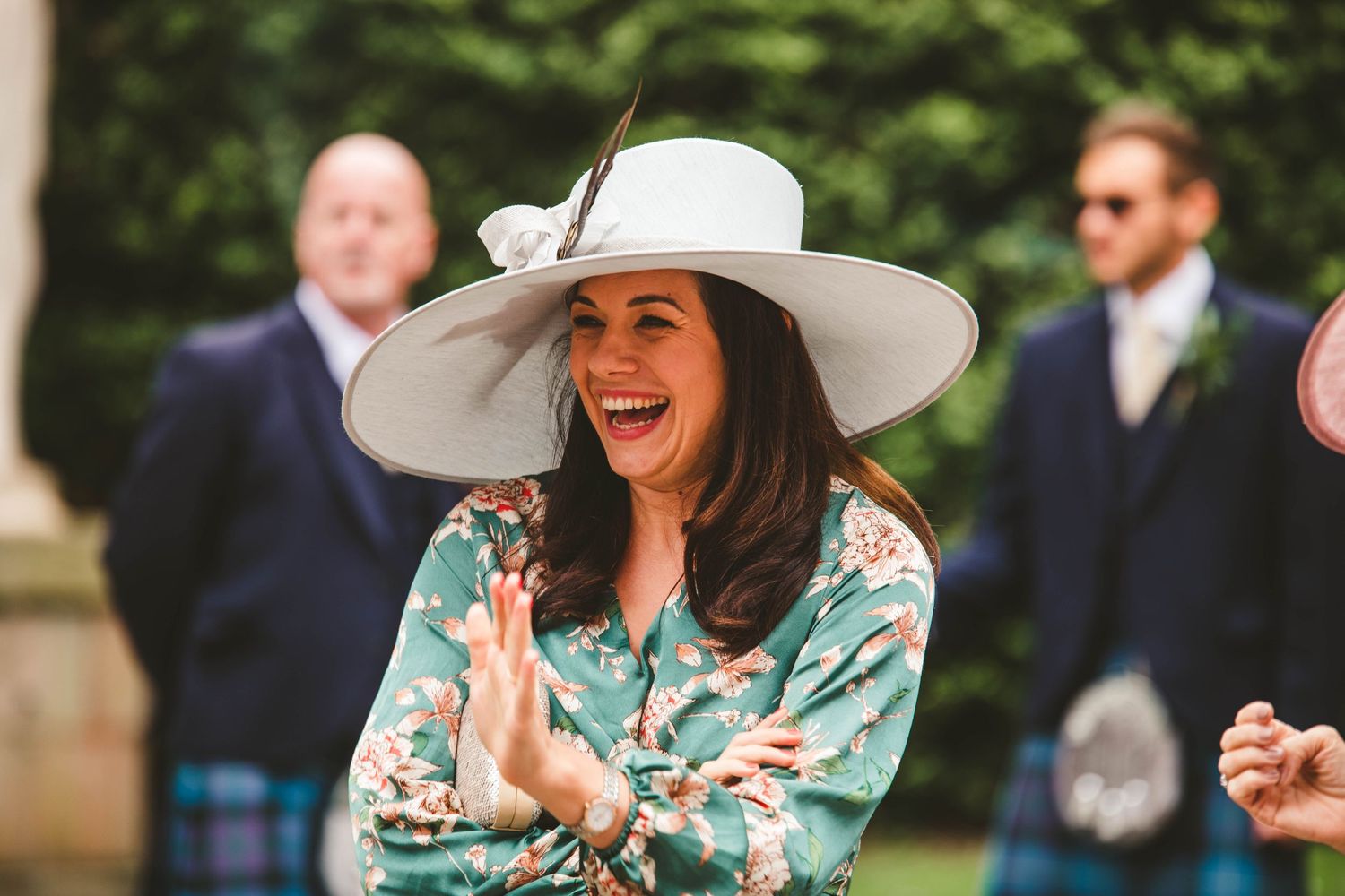 Guest in a wide-brimmed hat and floral dress laughing joyfully at an outdoor wedding.