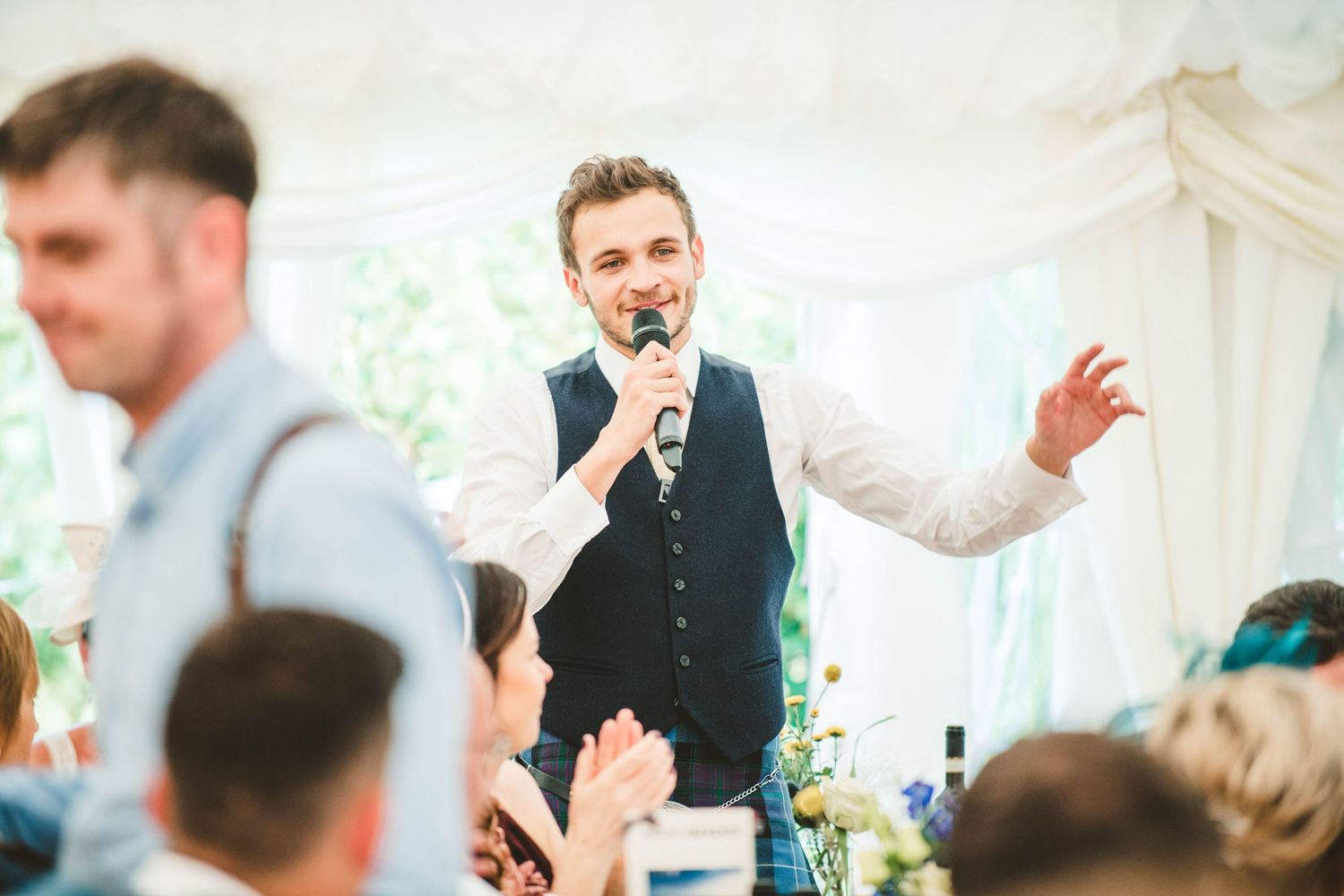 A wedding reception speech being delivered in a white marquee tent with guests seated at tables.