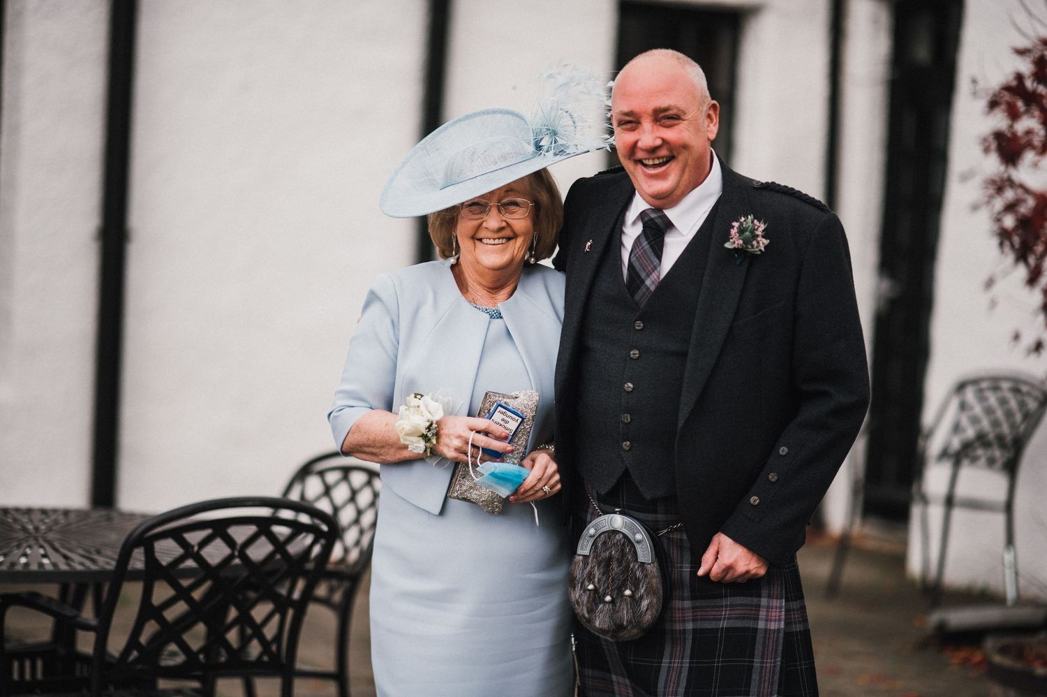 Wedding party members in formal attire and Scottish dress gather outside a white building.