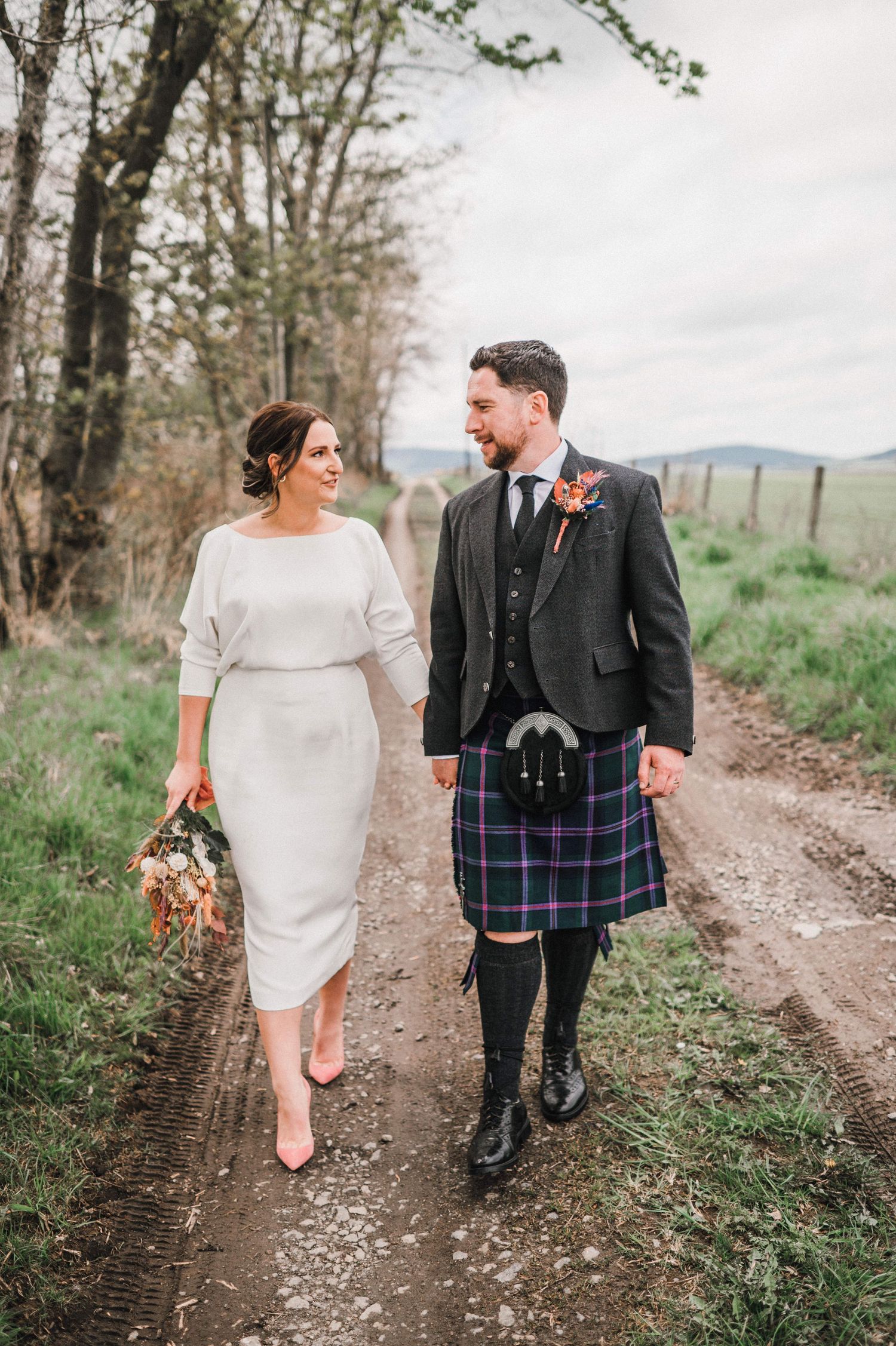 Elegant wedding couple walking along a rural path, groom in traditional Scottish attire.
