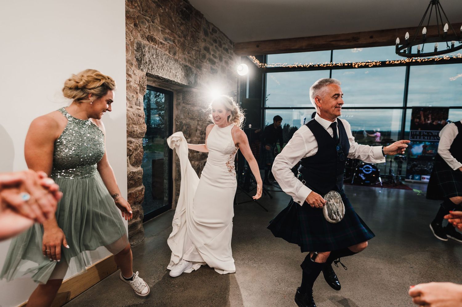 Wedding party dancing energetically in a modern venue with stone walls and large windows.
