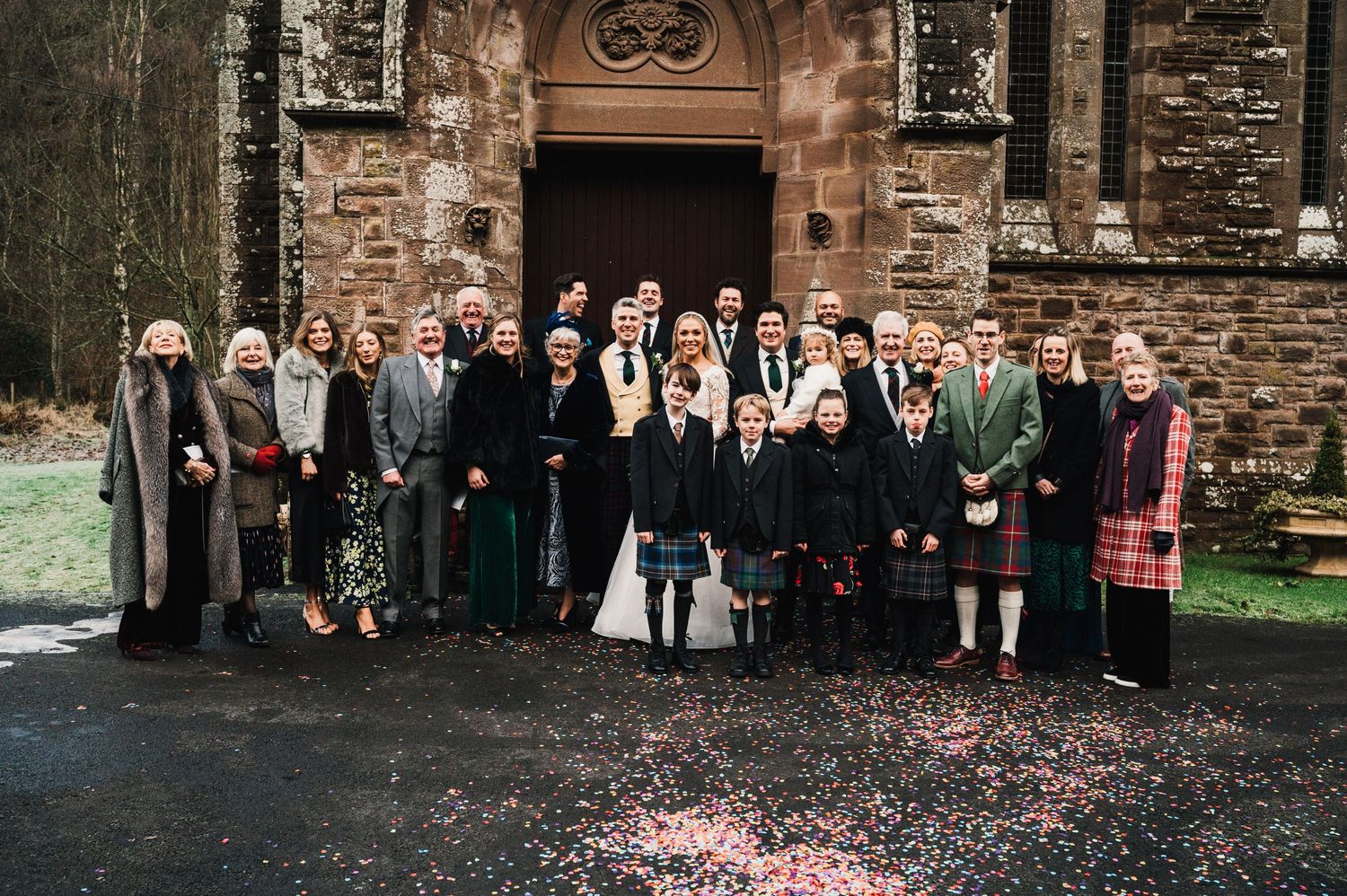 Wedding party poses for a formal group photo outside a stone church with scattered confetti on the ground.