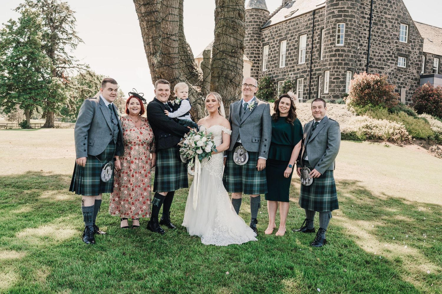 A wedding group photo with people wearing traditional Scottish kilts outside a stone church.