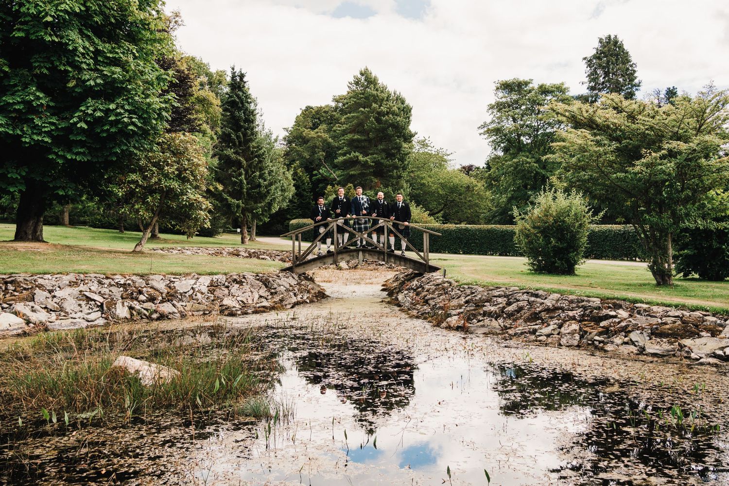 A scenic stone bridge reflects in a pond surrounded by lush gardens and trees.