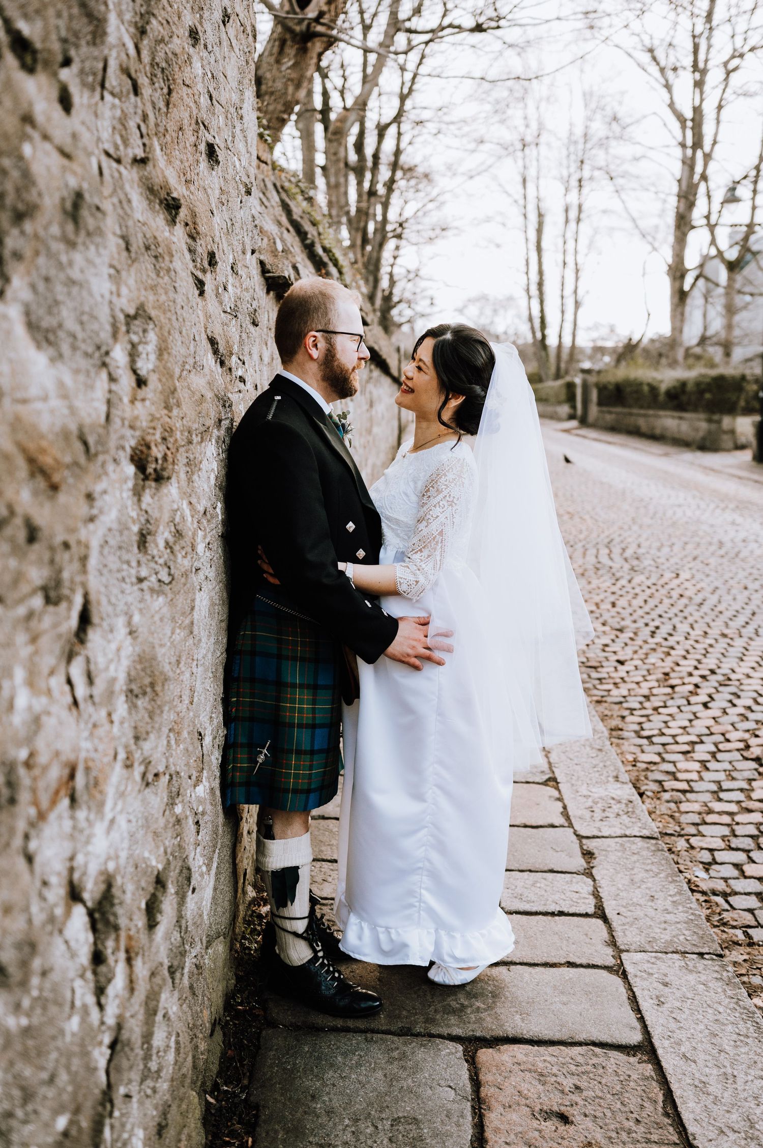 A romantic wedding couple in traditional Scottish attire pose against a stone wall on a cobblestone path.