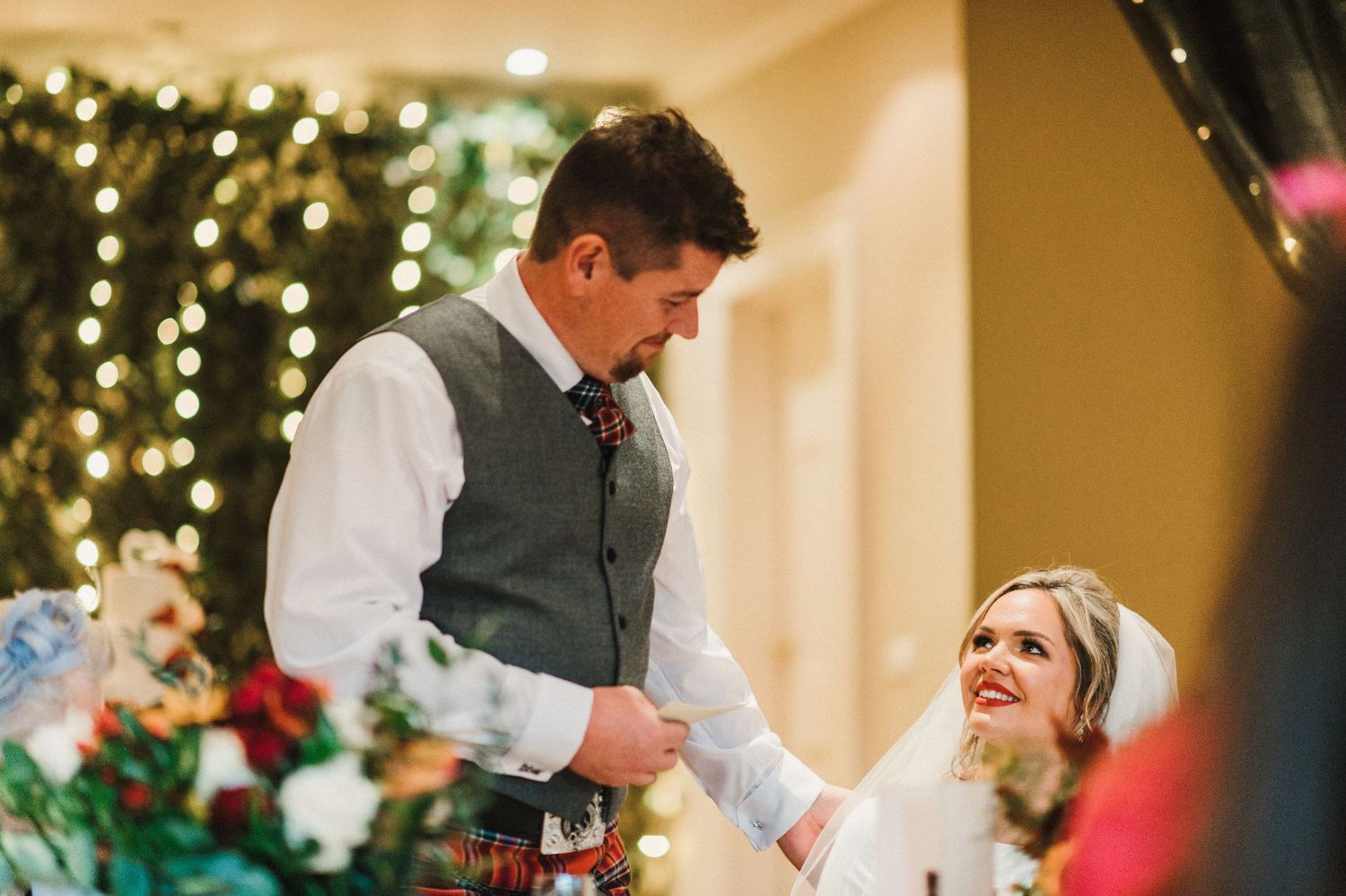 A wedding couple sharing a candid moment at their reception with twinkle lights and floral decor in the background.