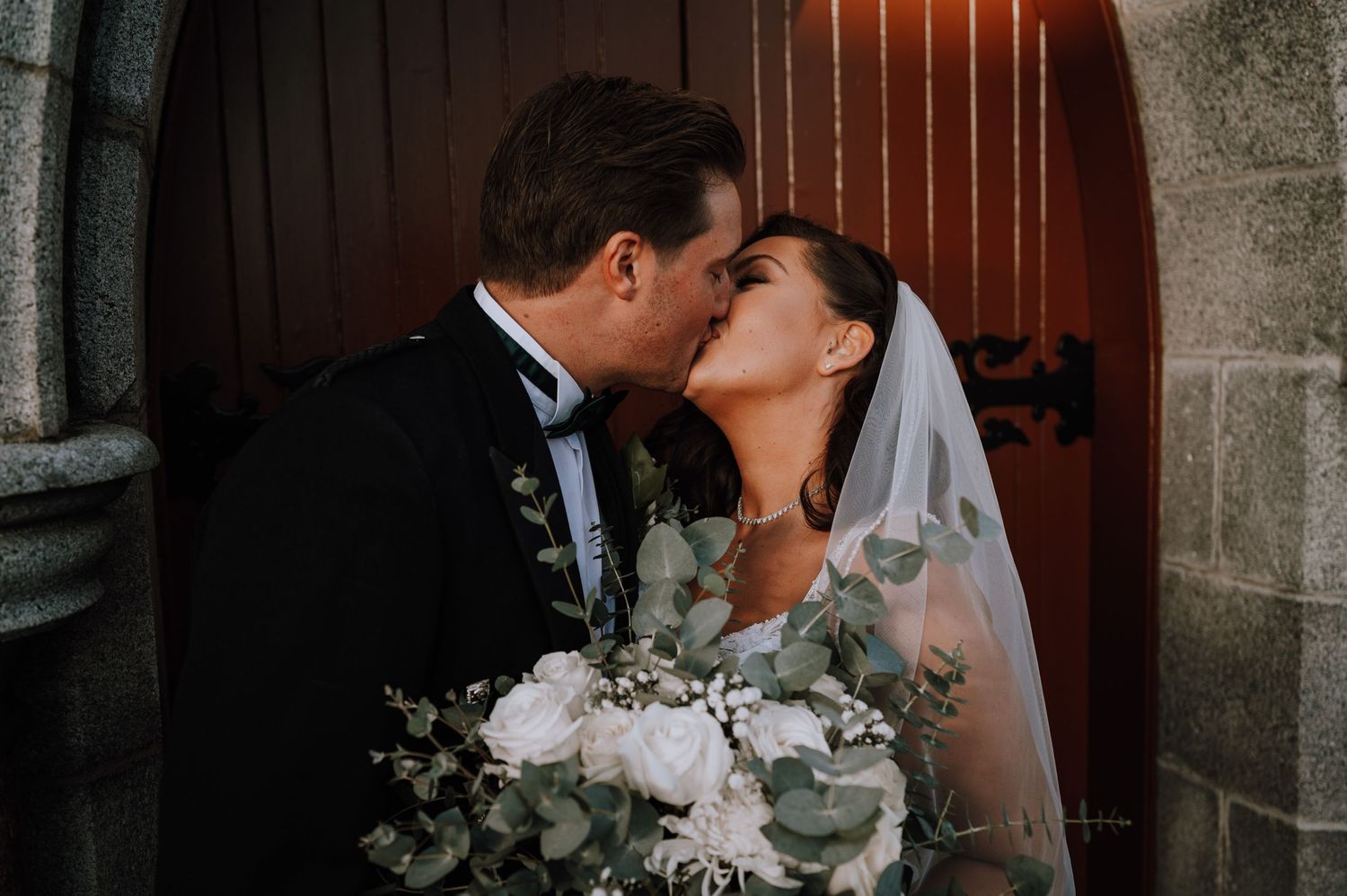 Romantic wedding couple sharing intimate kiss in stone church doorway.