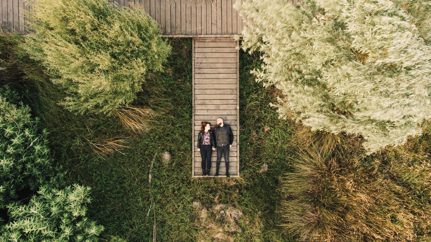 Drone shot of couple on wooden walkway surrounded by lush green trees.