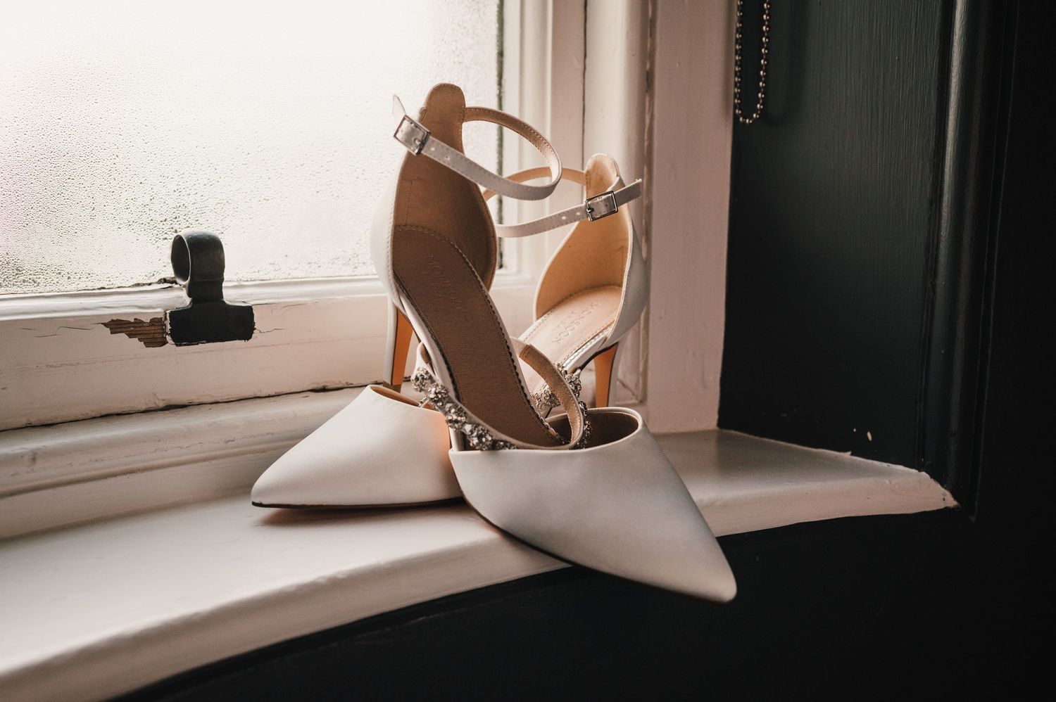 Elegant white pointed-toe wedding heels with ankle straps sitting on a windowsill.