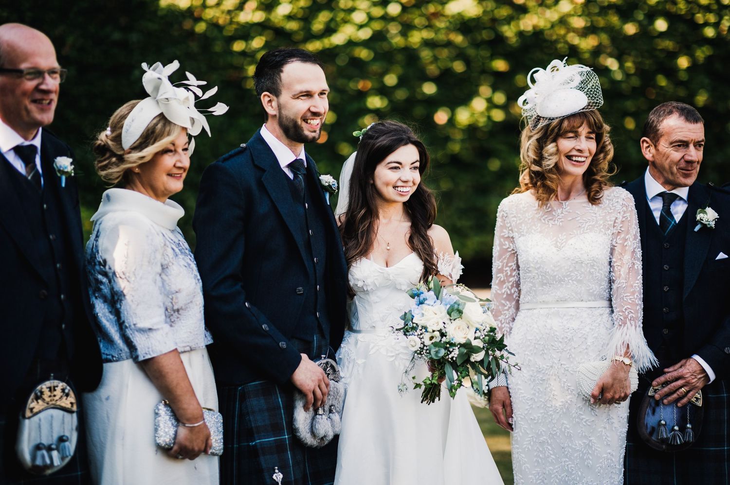 Wedding party gathered outdoors with elegant attire including traditional Scottish kilts and formal dresses.