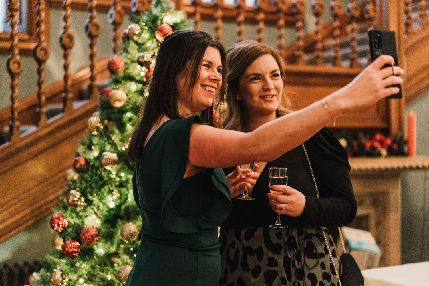 Two women taking a selfie by a decorated Christmas staircase with wine glasses in hand.