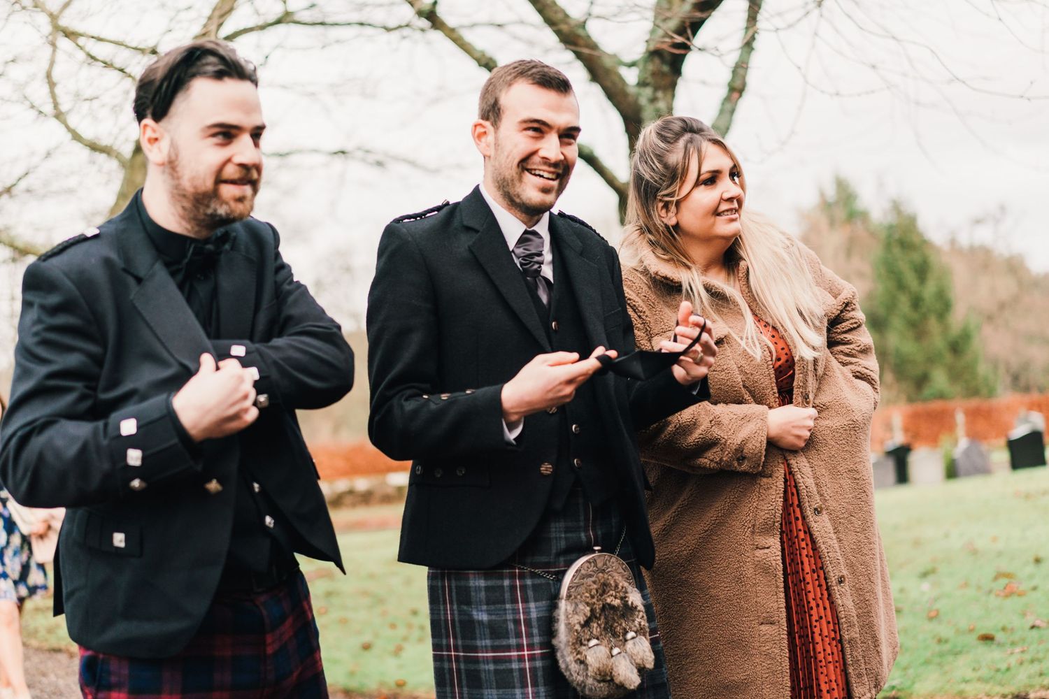 Wedding guests in traditional Scottish attire laughing together outdoors.