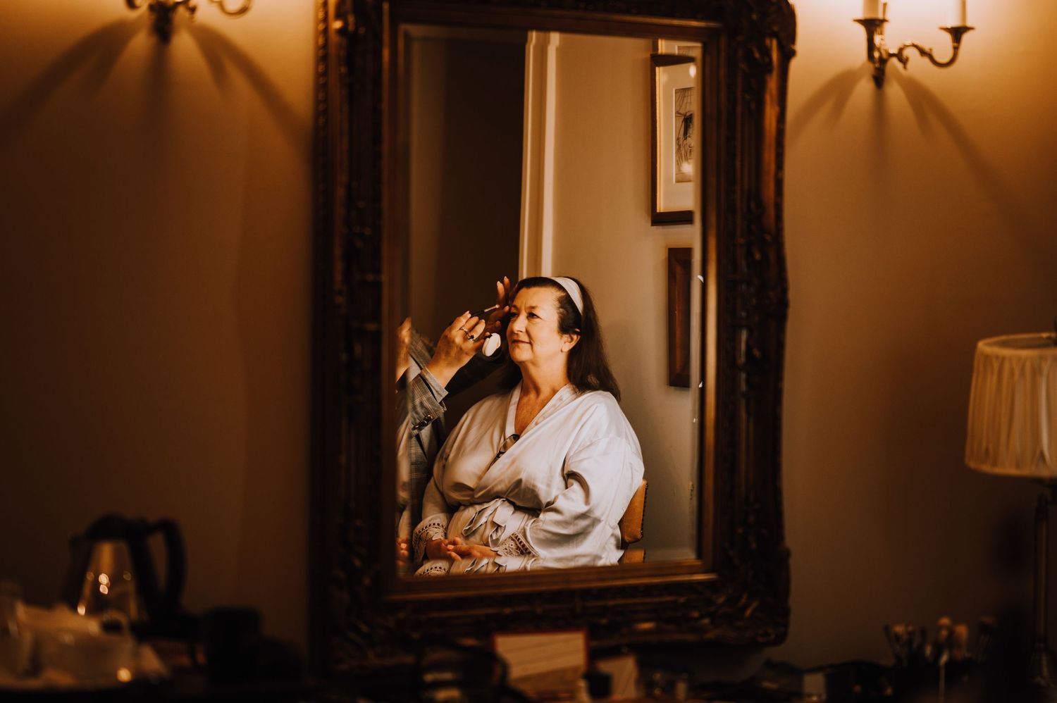 A reflection in an ornate gold mirror shows someone applying makeup in warm lighting.