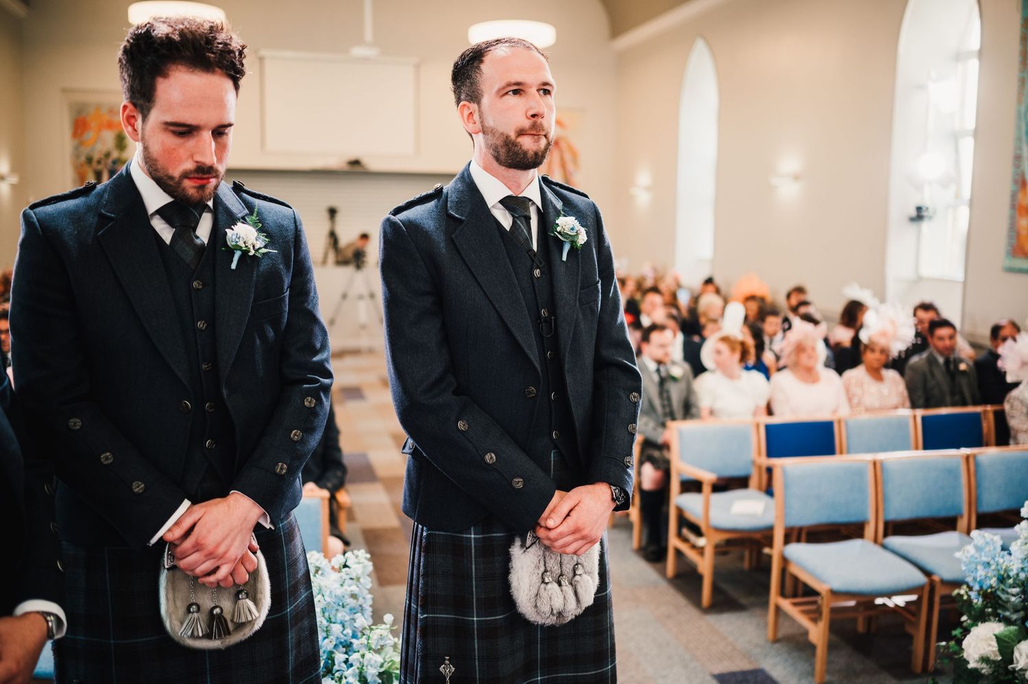 Two groomsmen in kilts and suits stand solemnly during church ceremony.