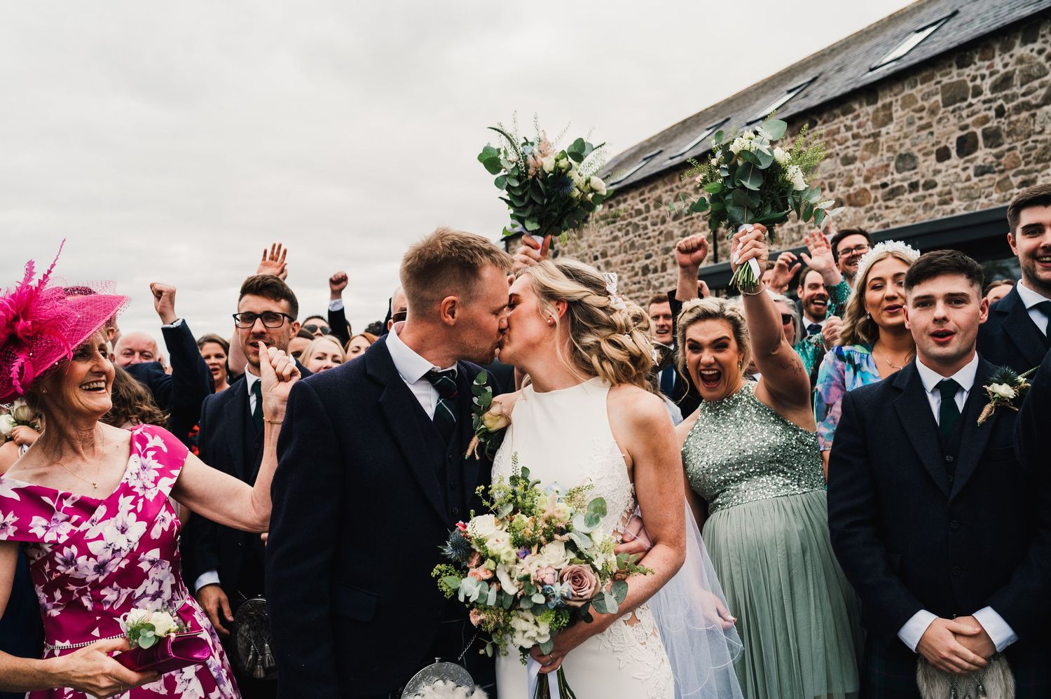Wedding couple shares romantic kiss surrounded by cheering guests outside stone venue.