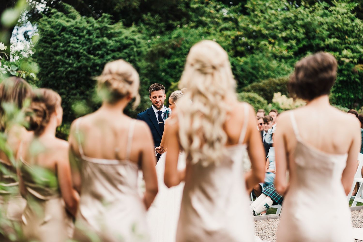 Bridesmaids in pale dresses seen from behind during outdoor garden ceremony.
