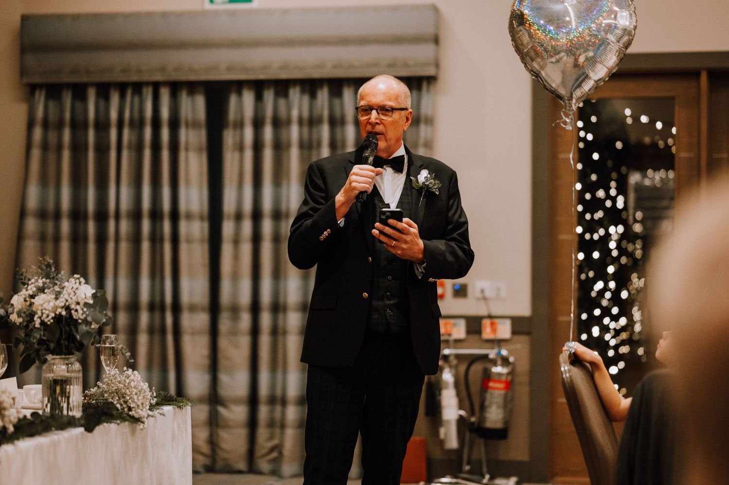 Speaker in formal attire delivers speech at wedding reception with string lights backdrop.