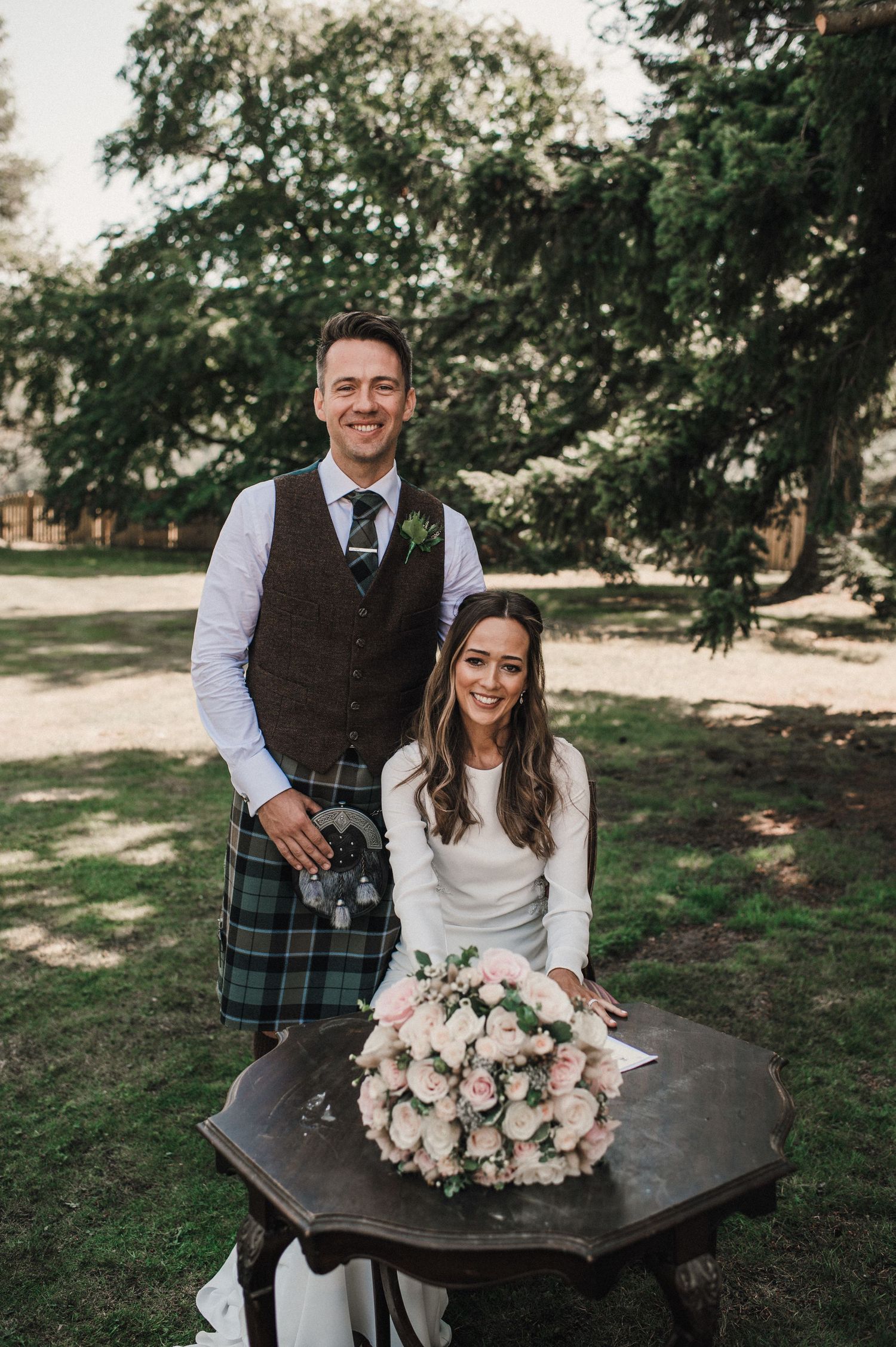 A couple in wedding attire poses with a stunning pink and white floral bouquet in an outdoor garden setting.