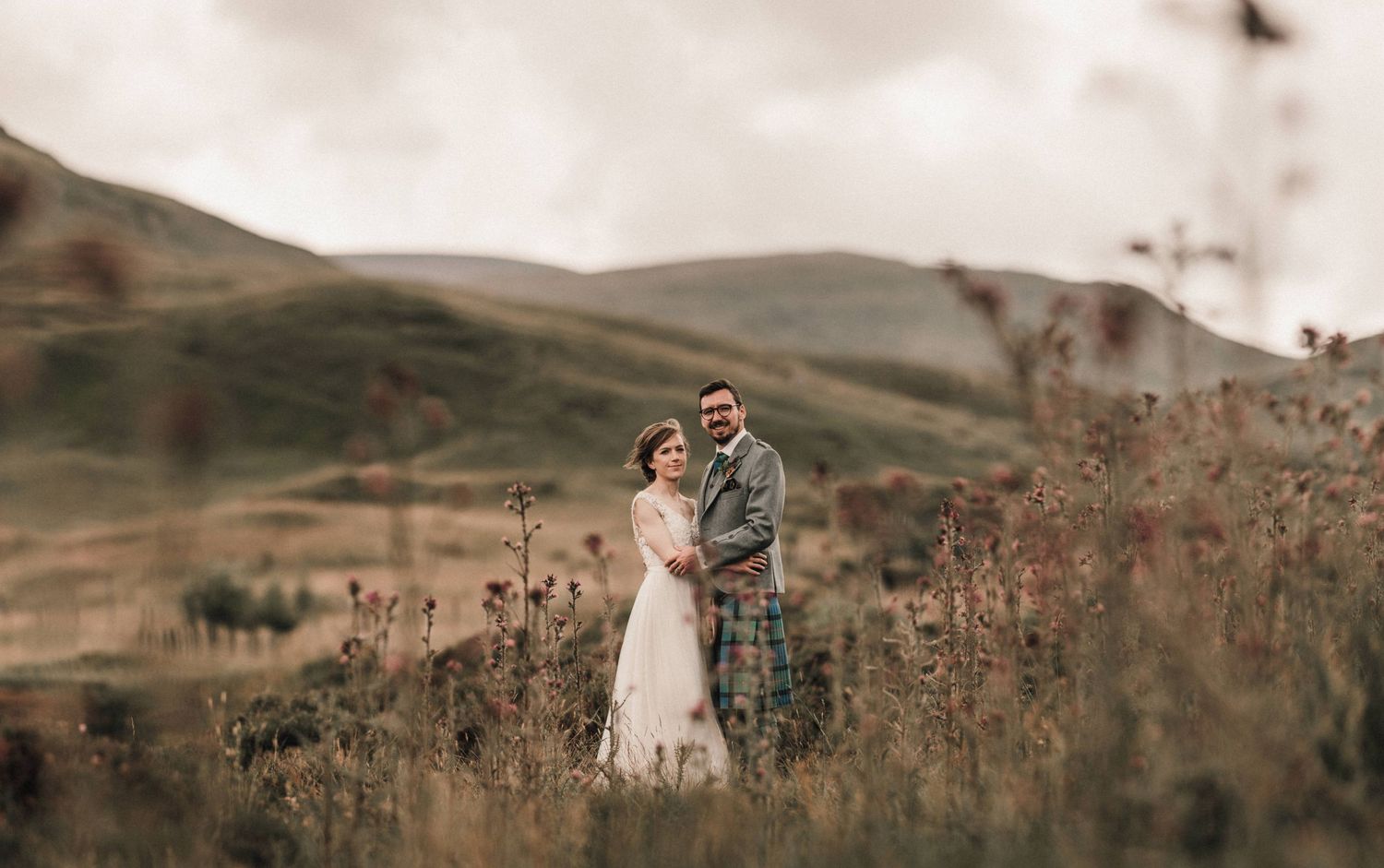Wedding couple standing in tall grass with misty Scottish mountains in the background.