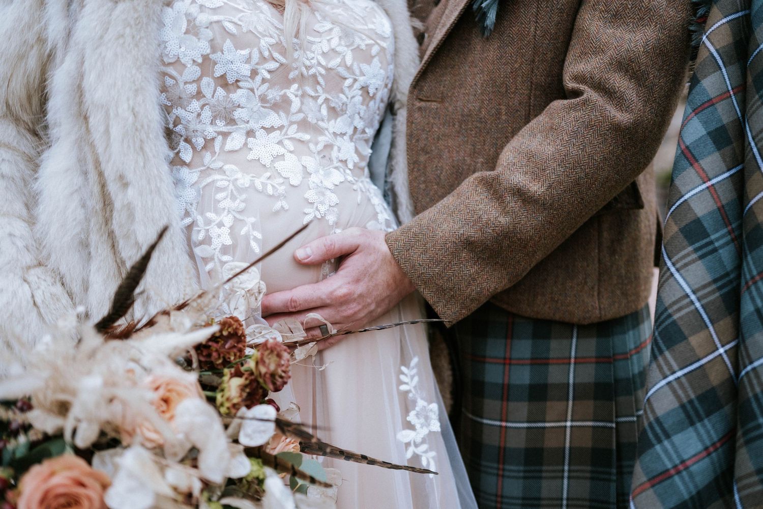 Close-up detail of wedding attire showing white lace dress and brown tweed jacket with tartan pattern.