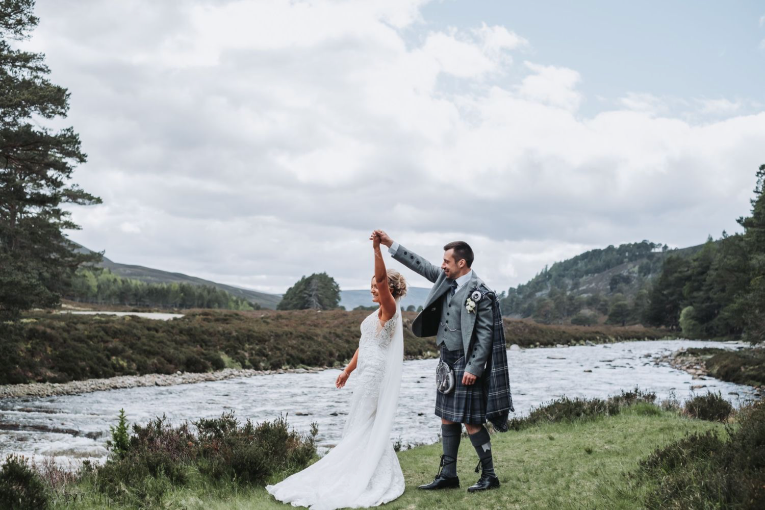 A couple in wedding attire dance together by a river in the Scottish Highlands, with mountains in the background.