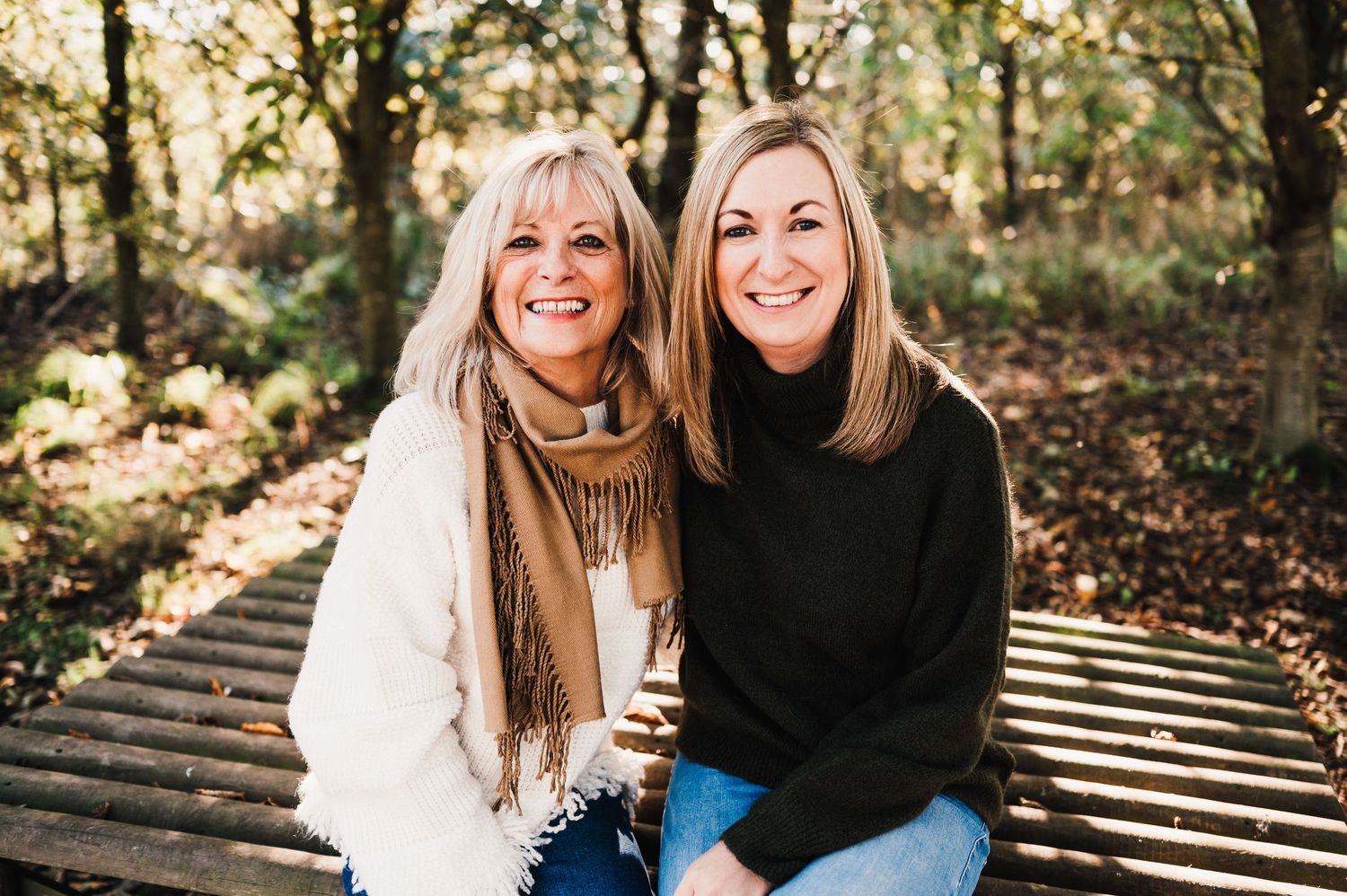 Two people sit together on wooden steps surrounded by autumn leaves in a sunlit forest setting.