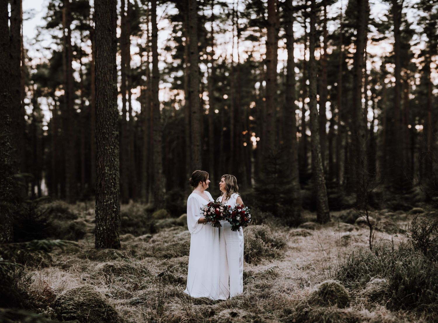 Same Sex Wedding Photography. Two people in white dresses hold bouquets in moody forest setting at dusk.