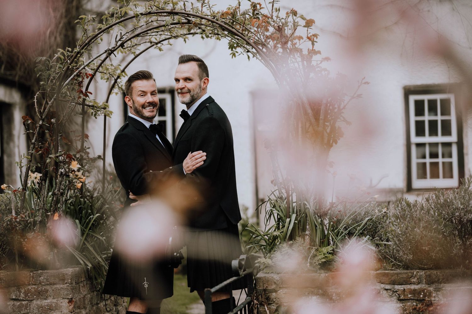 Logie House Wedding Photography. Two people in black tuxedos share an intimate moment in a rustic outdoor setting.