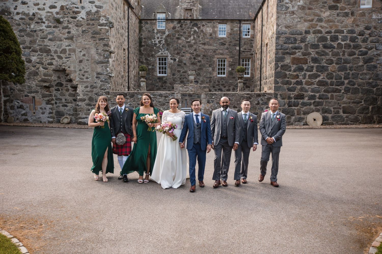 Wedding party group photo in front of a historic stone building with rustic architecture.