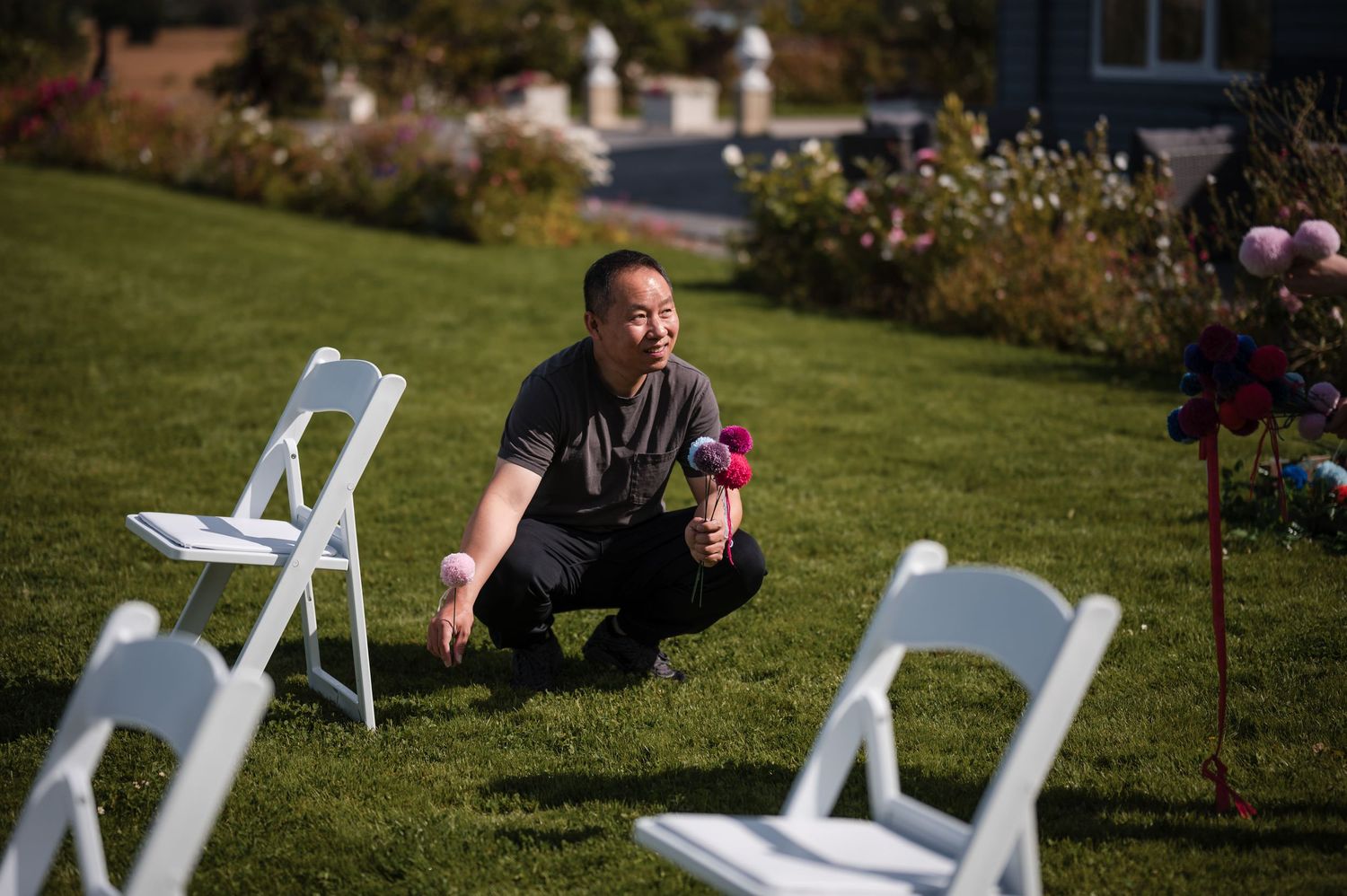A person sits cross-legged on grass between white folding chairs in a garden setting.