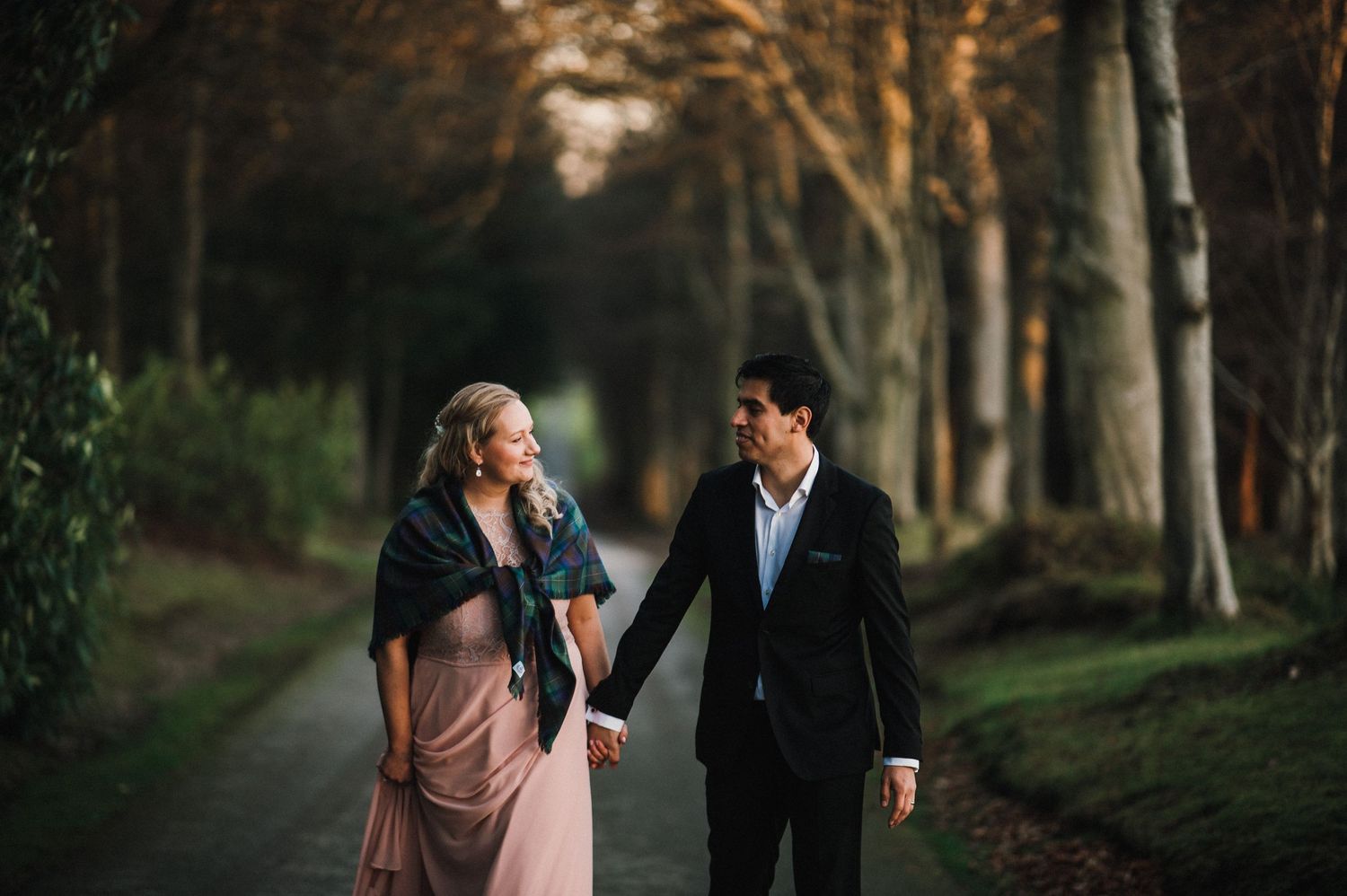 A couple walks hand in hand along a tree-lined path at dusk wearing formal attire.