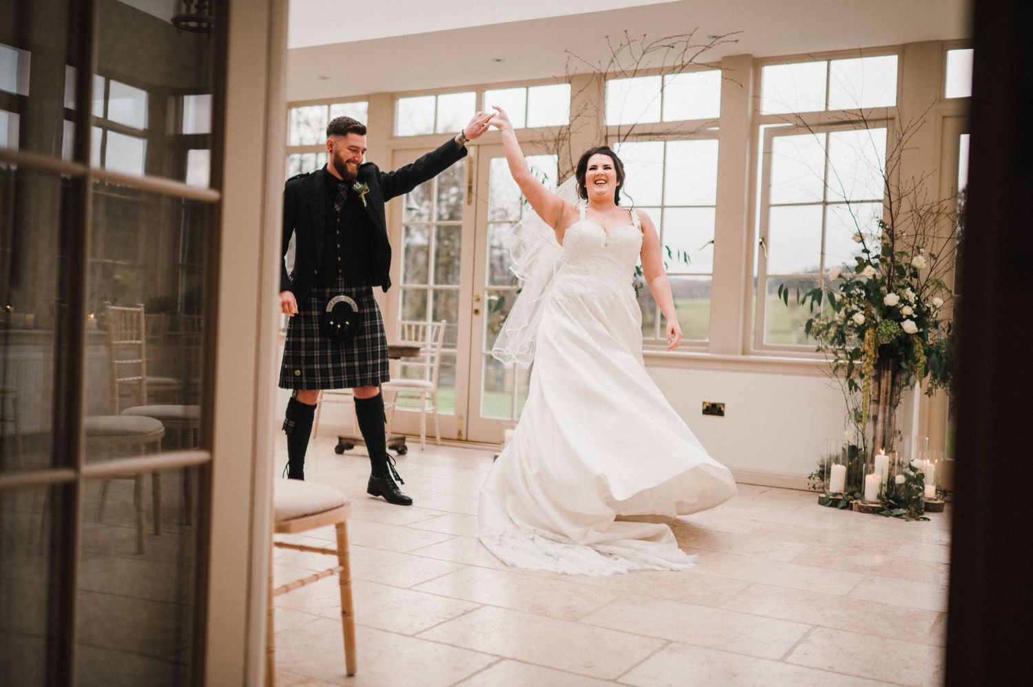 Couple in wedding attire dance and twirl in bright conservatory setting.