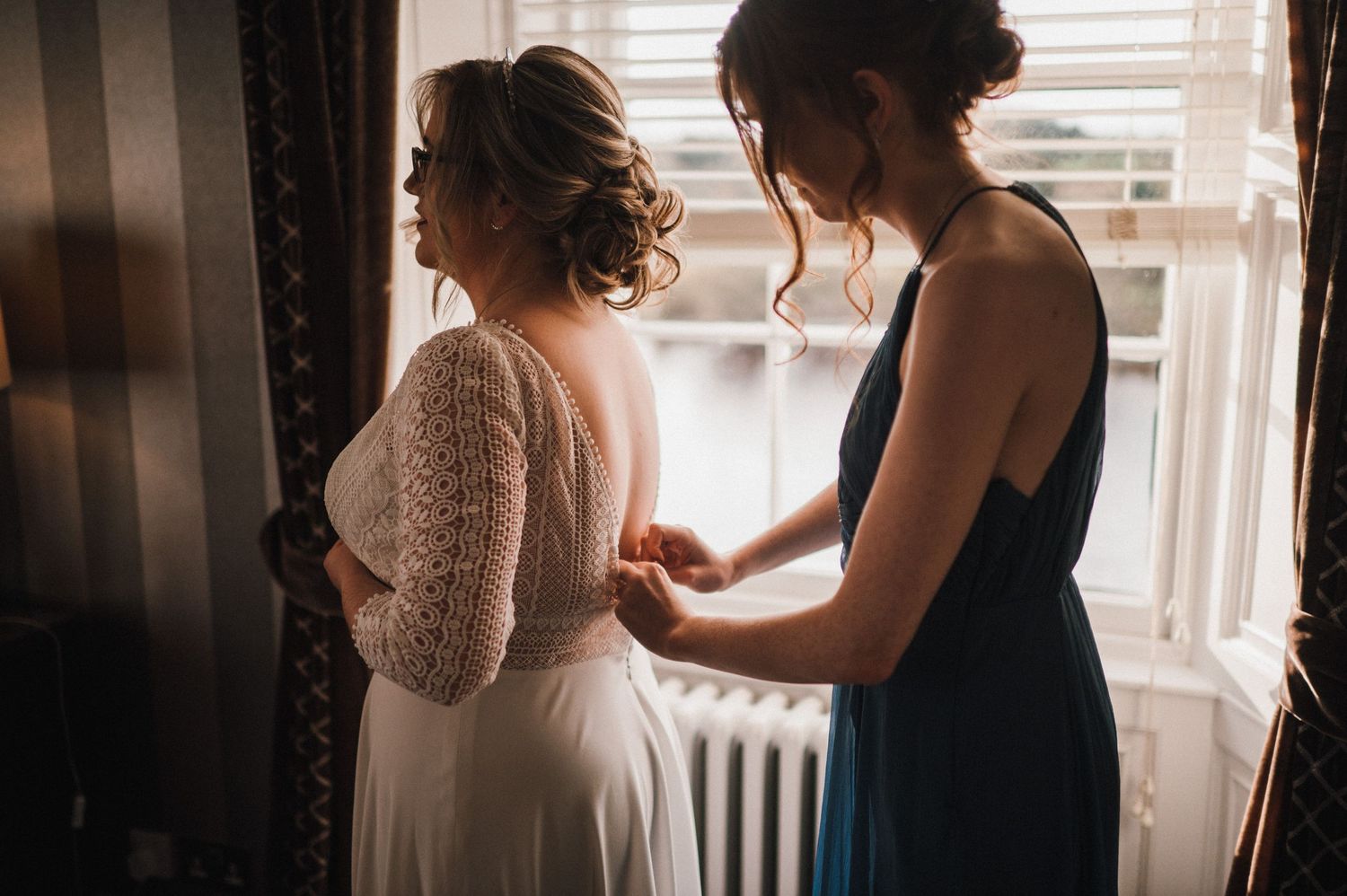 Two figures in a sunlit room preparing wedding attire near a window.