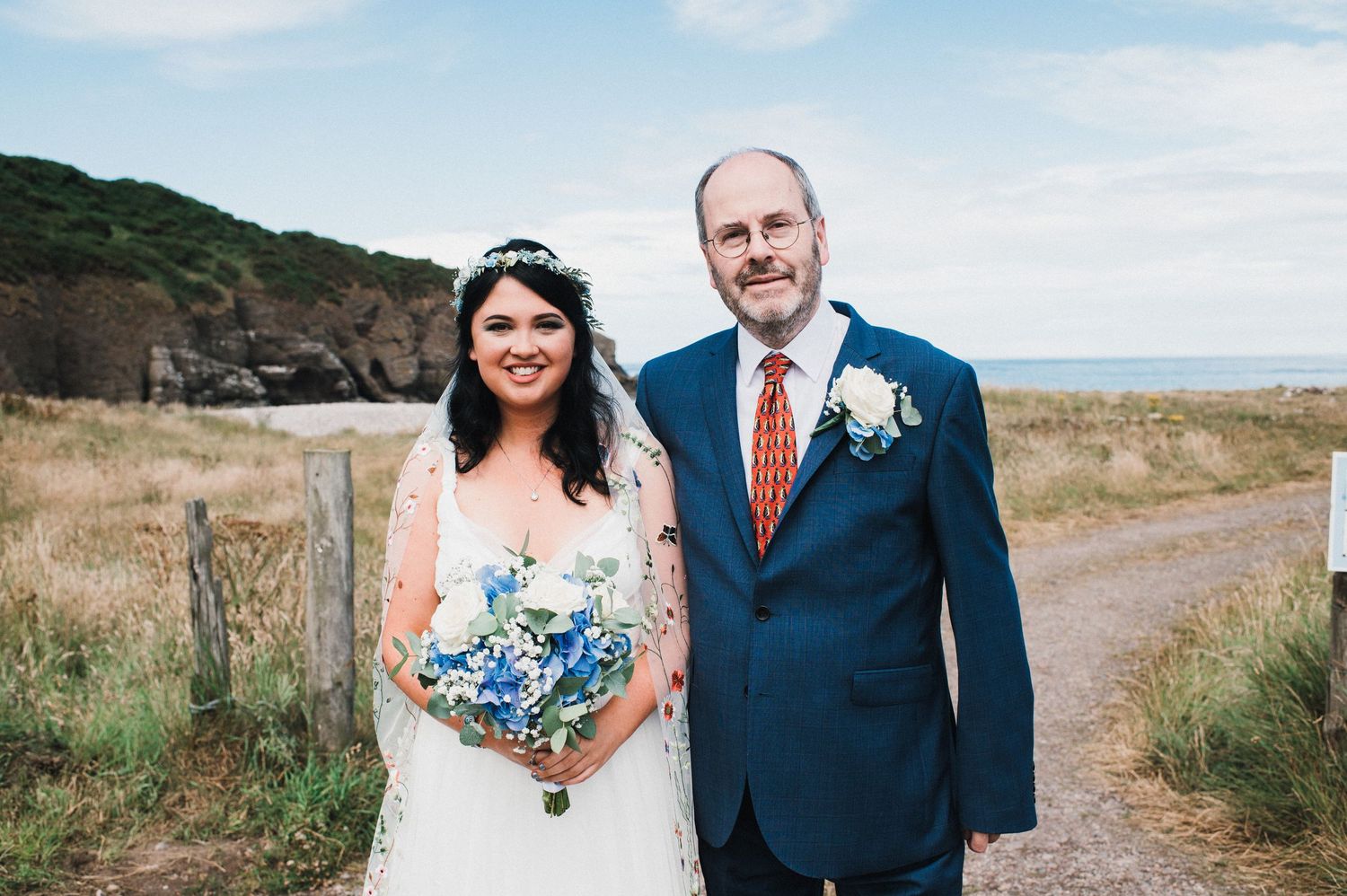 Newlyweds pose together on coastal path with ocean cliffs in background.