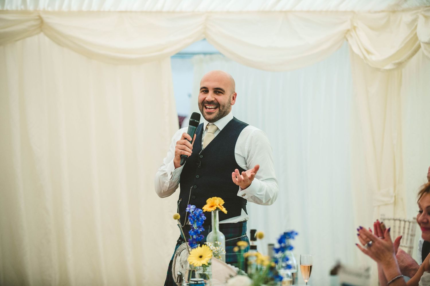 Speaker in waistcoat delivering speech at wedding reception with yellow flowers on table.