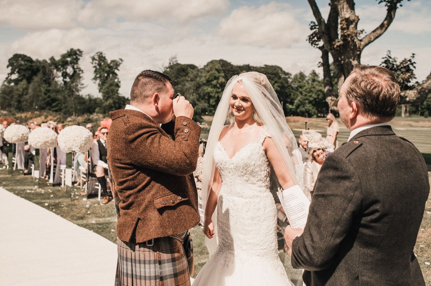 A touching wedding moment in front of gathered guests at an outdoor ceremony.
