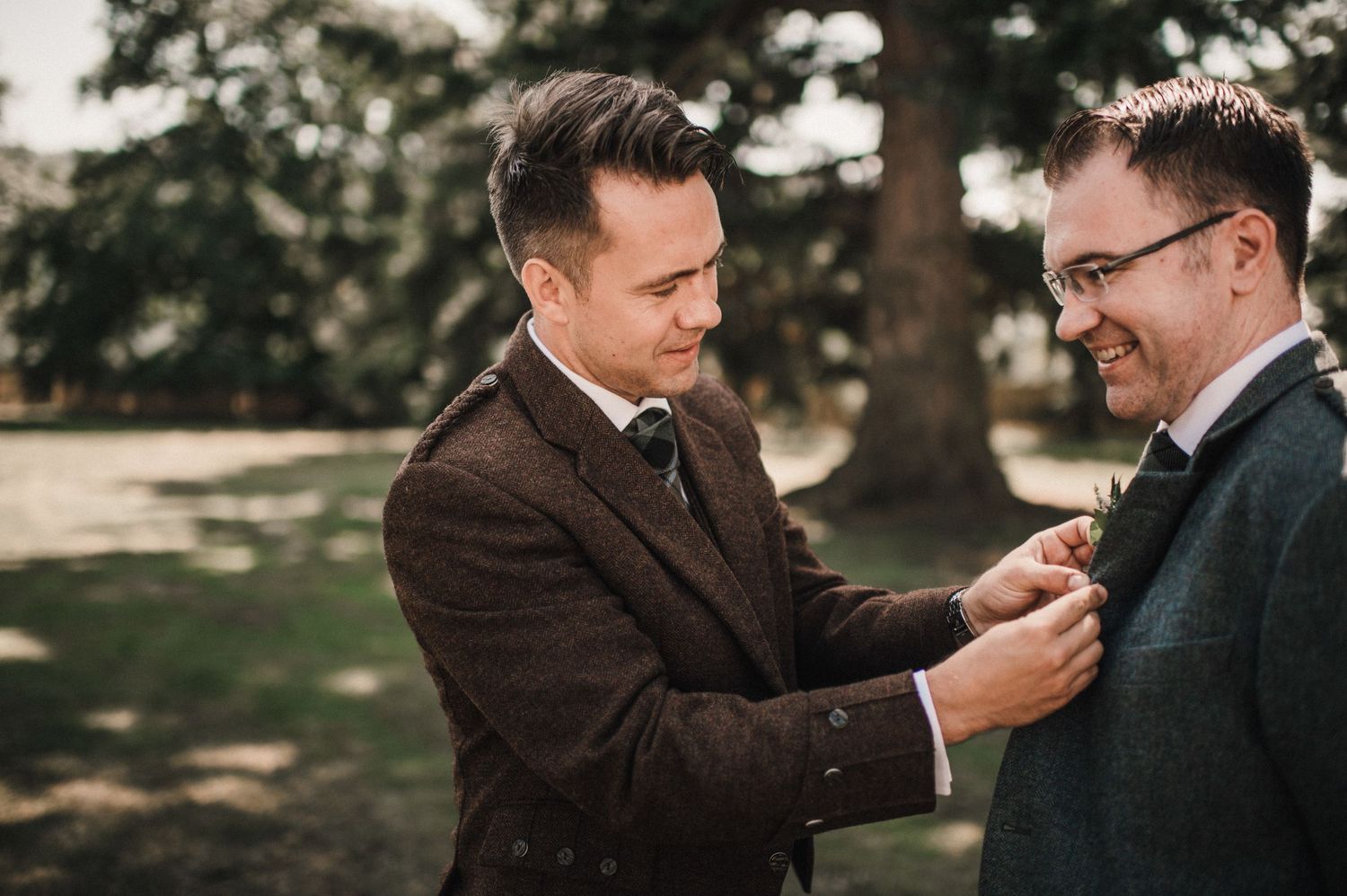 Two people in formal suits adjusting boutonnieres outdoors.