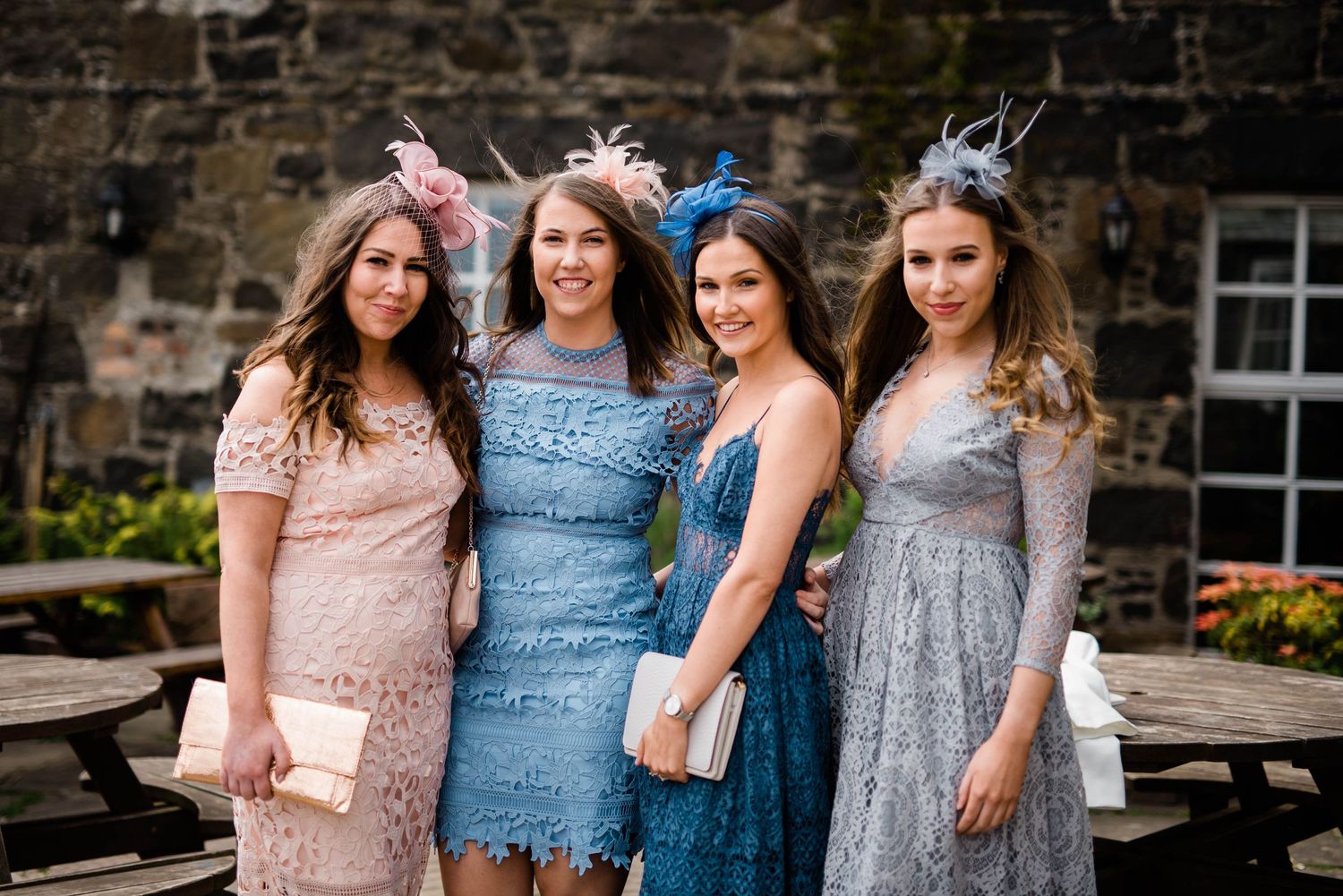 Group of wedding guests in pastel lace dresses posing against stone wall.