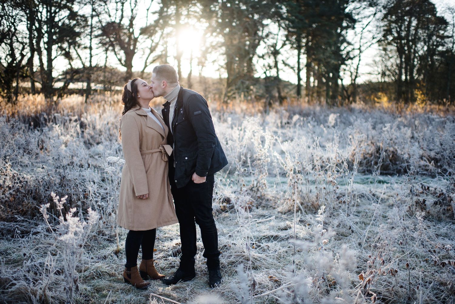 A couple shares an intimate moment in a snow-dusted winter field at sunset.