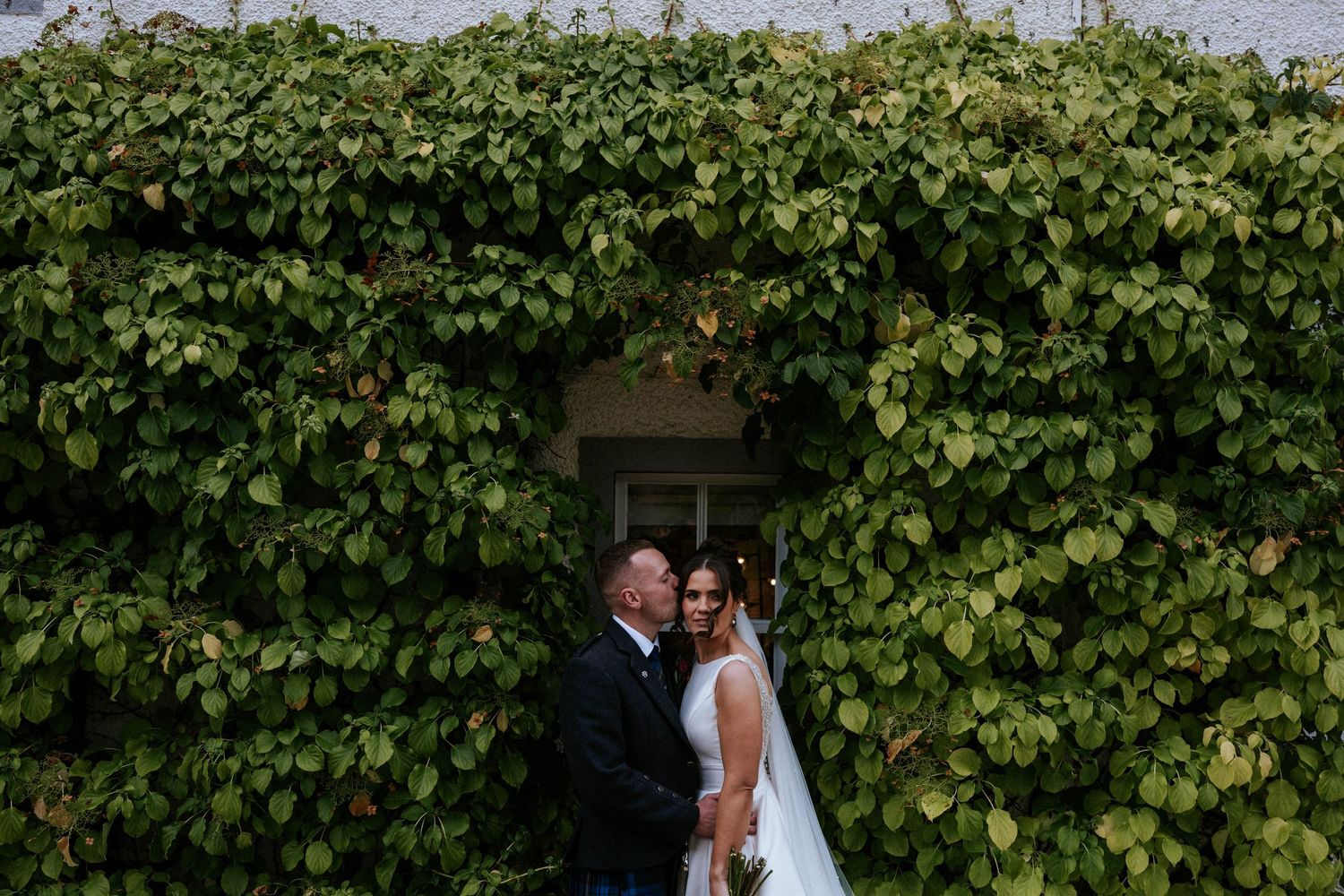 A couple poses in front of an ivy-covered wall during their wedding celebration.