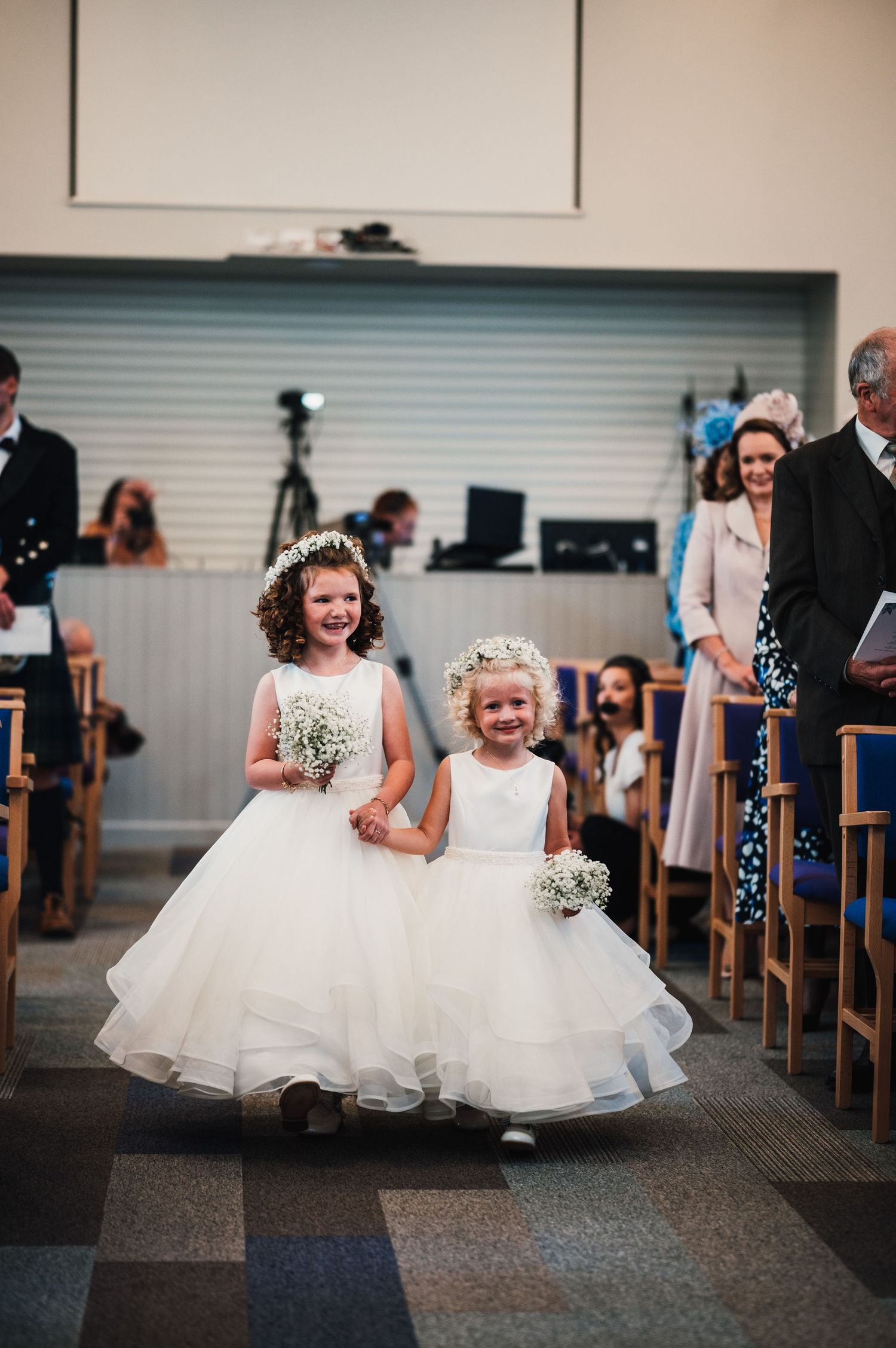 Two young flower girls in white tulle dresses smile and walk together holding small bouquets in a church aisle.