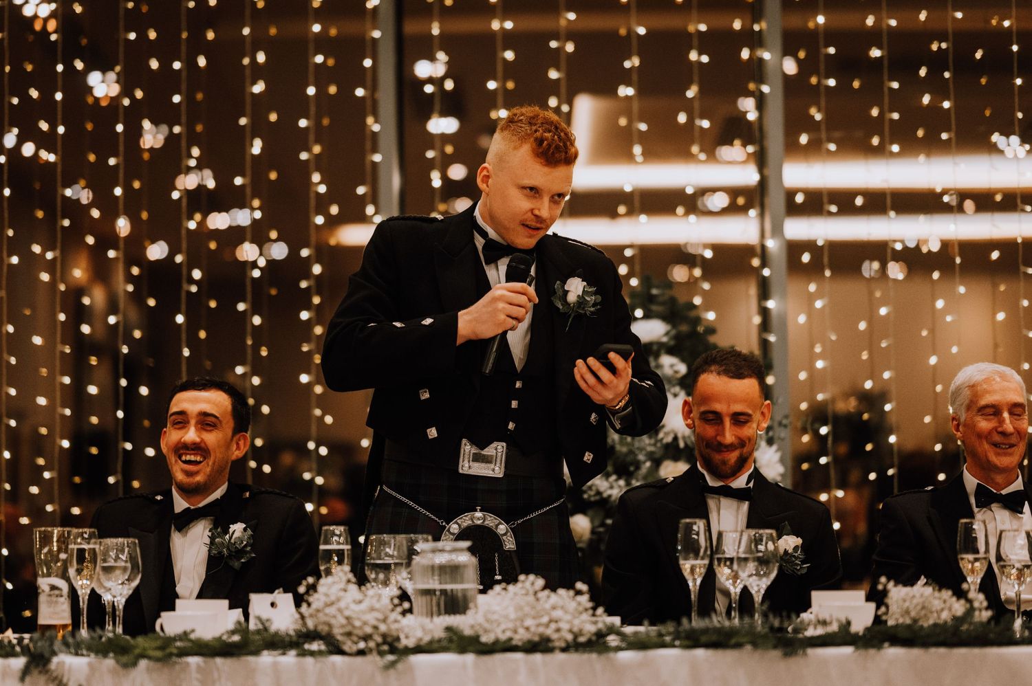 Speaker in formal attire addresses guests at elegant wedding reception with twinkle light backdrop.