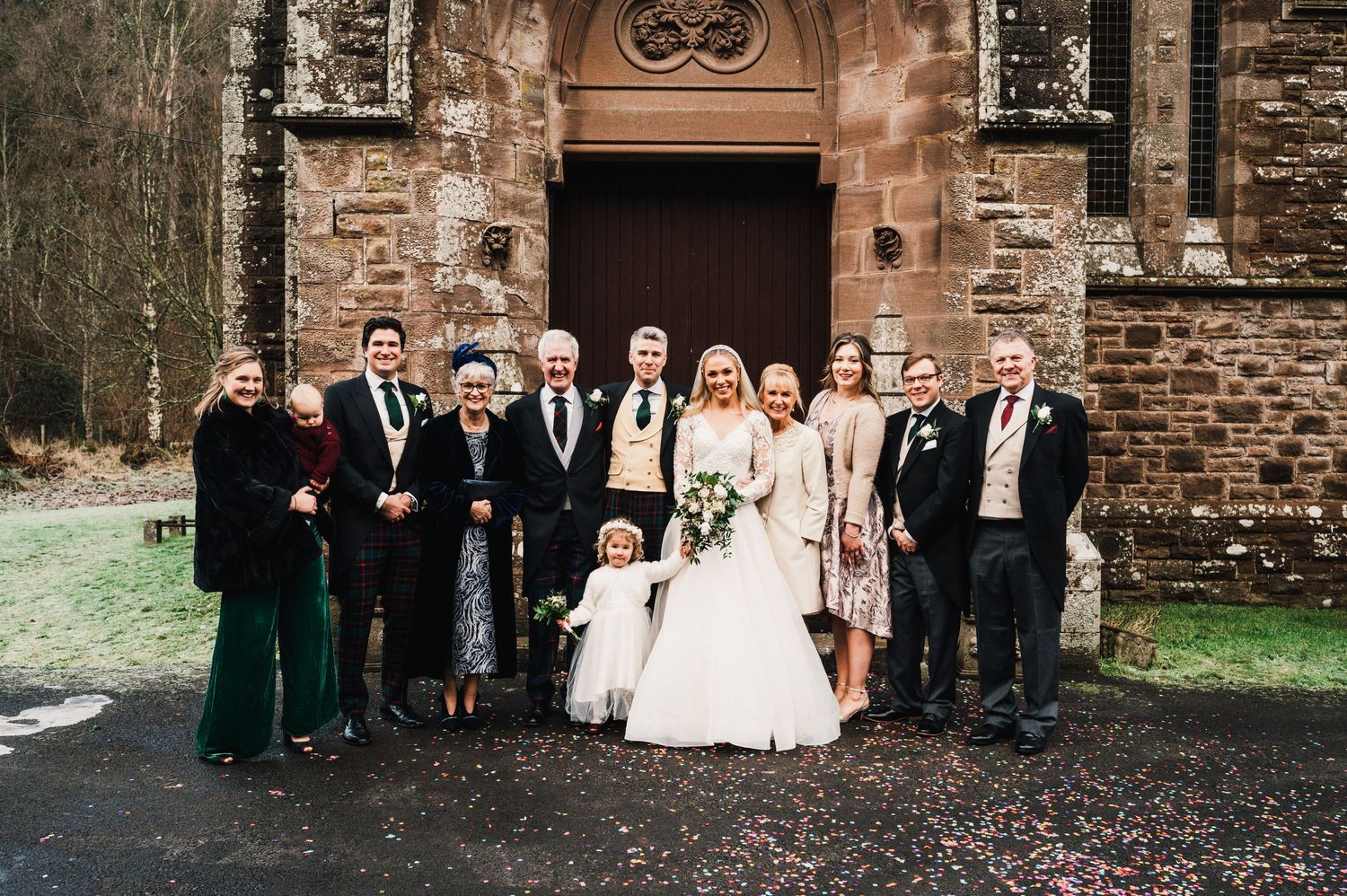 Wedding party poses in front of historic stone church with dark wooden door and red brick facade.