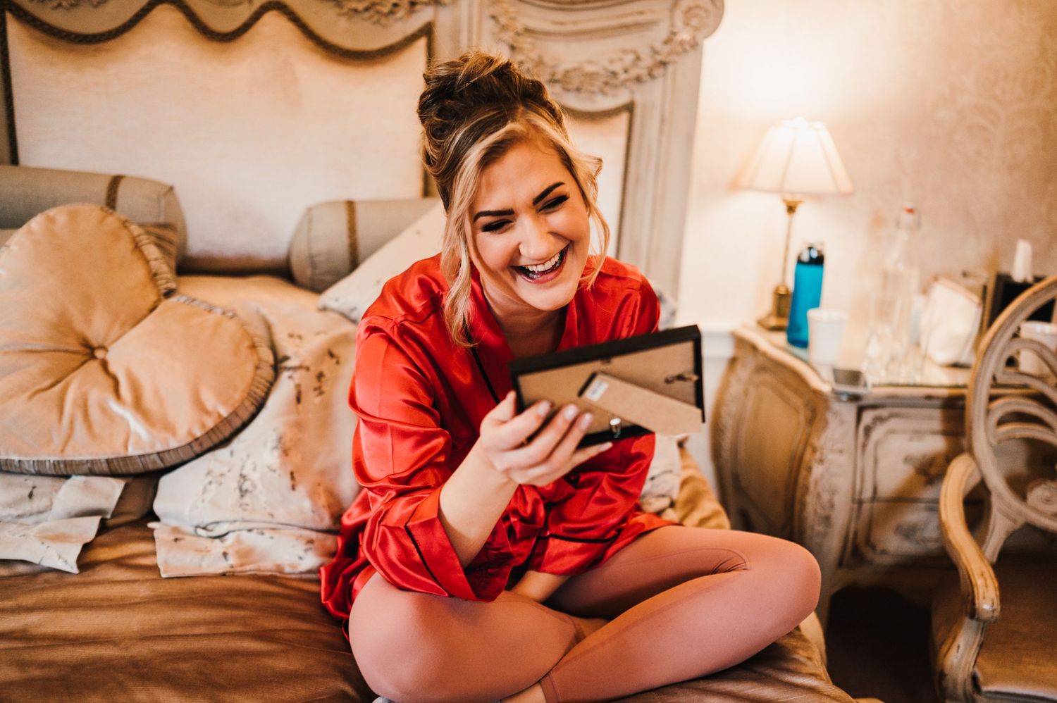Person in red silk robe smiling while looking at tablet device in elegant bedroom setting.