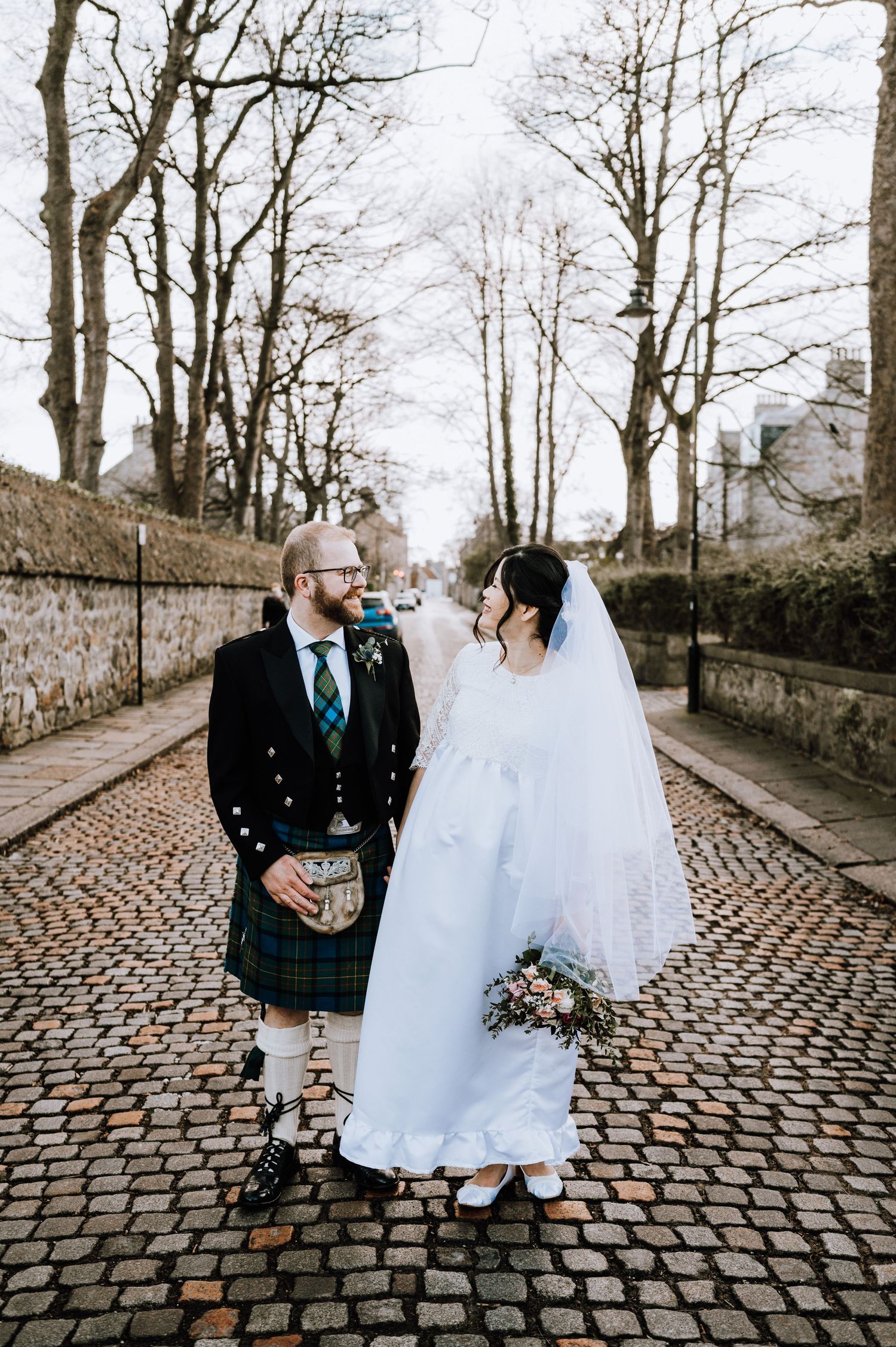 A wedding couple poses on cobblestone street, groom in traditional Scottish kilt and bride in elegant white gown.